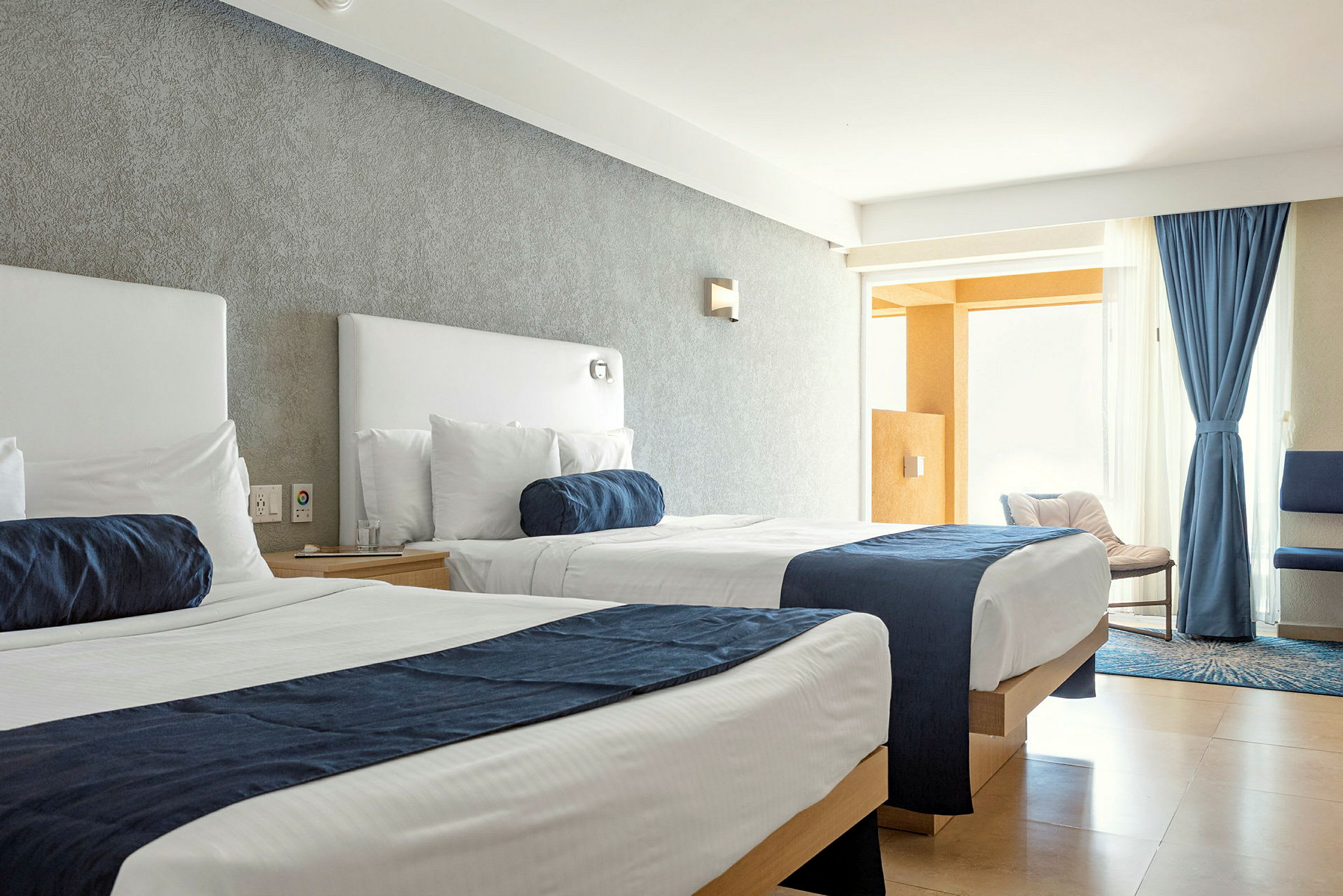 Hotellihuone, jossa kaksi sänkyä, joissa valkoiset lakanat ja siniset päiväpeitteet. Ikkunan siniset verhot päästävät valoa huoneeseen. (Tekoälyn luoma teksti)
