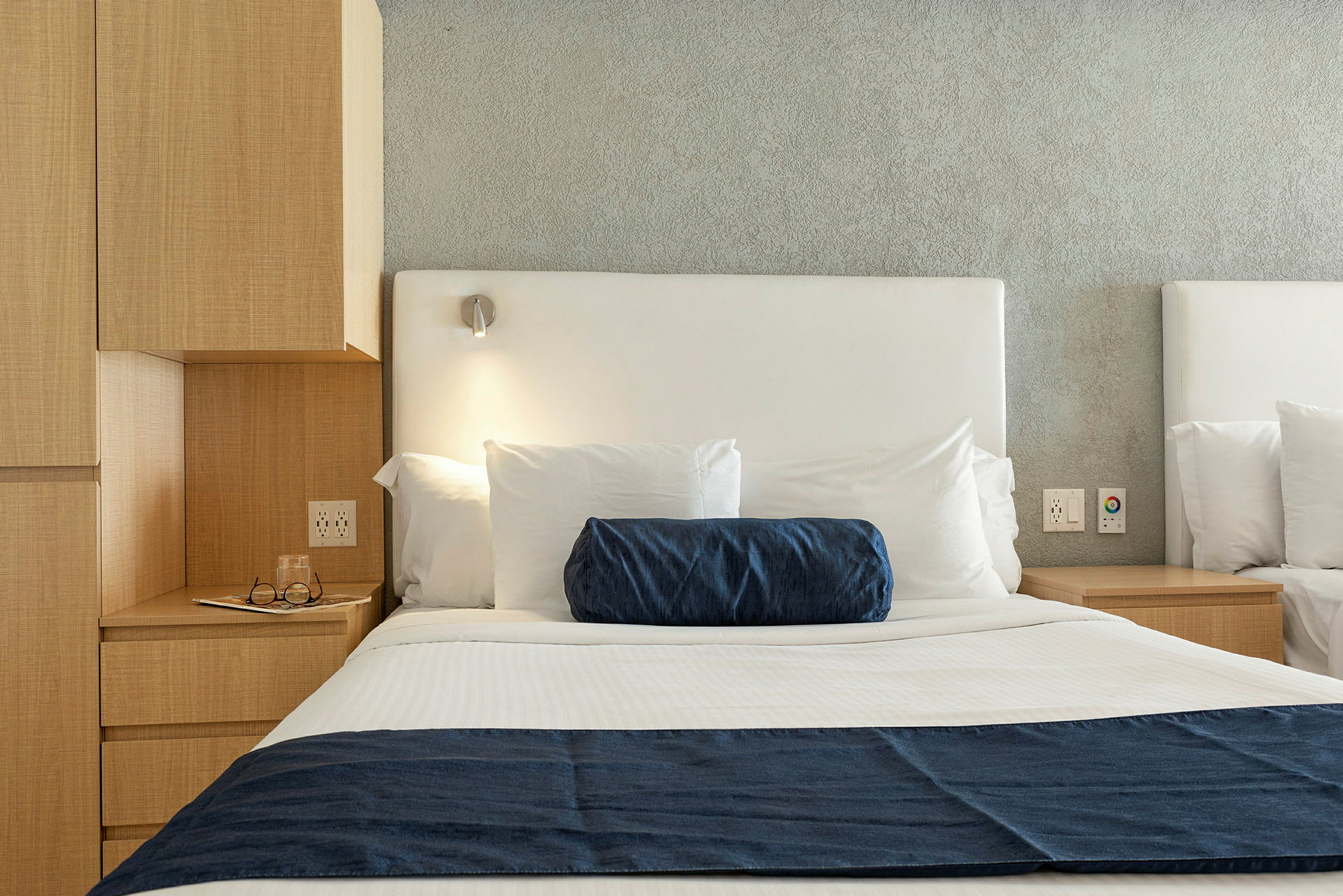 Kuva hotellisängystä, jossa on valkoiset lakanat, sininen koristetyyny ja sininen sängynpeite sekä yöpöydän lamppu. (Tekoälyn luoma teksti)