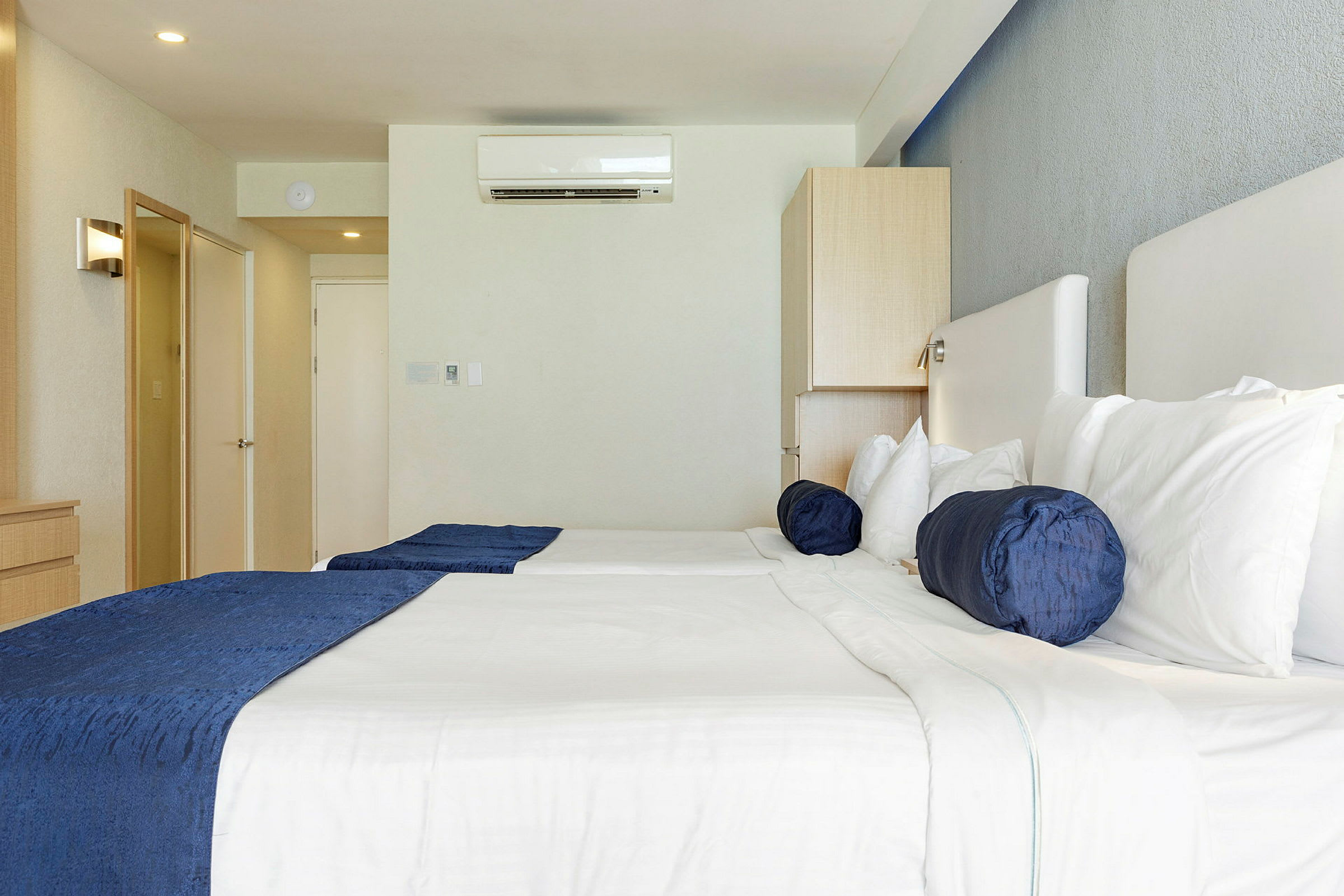 Hotellihuone, jossa kaksi sänkyä, valkoiset vuodevaatteet ja siniset tyynyt. Seinällä on ilmastointilaite. (Tekoälyn luoma teksti)