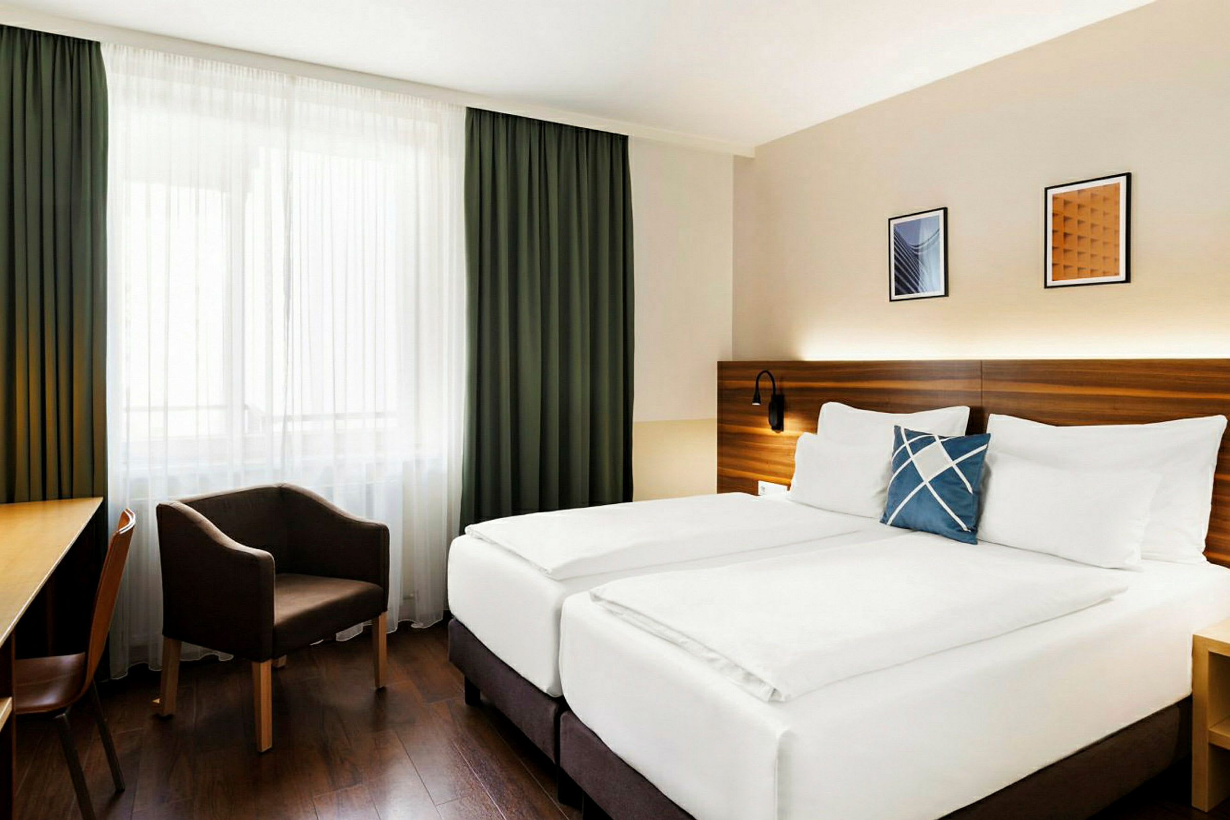 Hotellihuone, jossa on parisänky, nojatuoli ja kirjoituspöytä. Ikkunassa on verhot. (Tekoälyn luoma teksti)