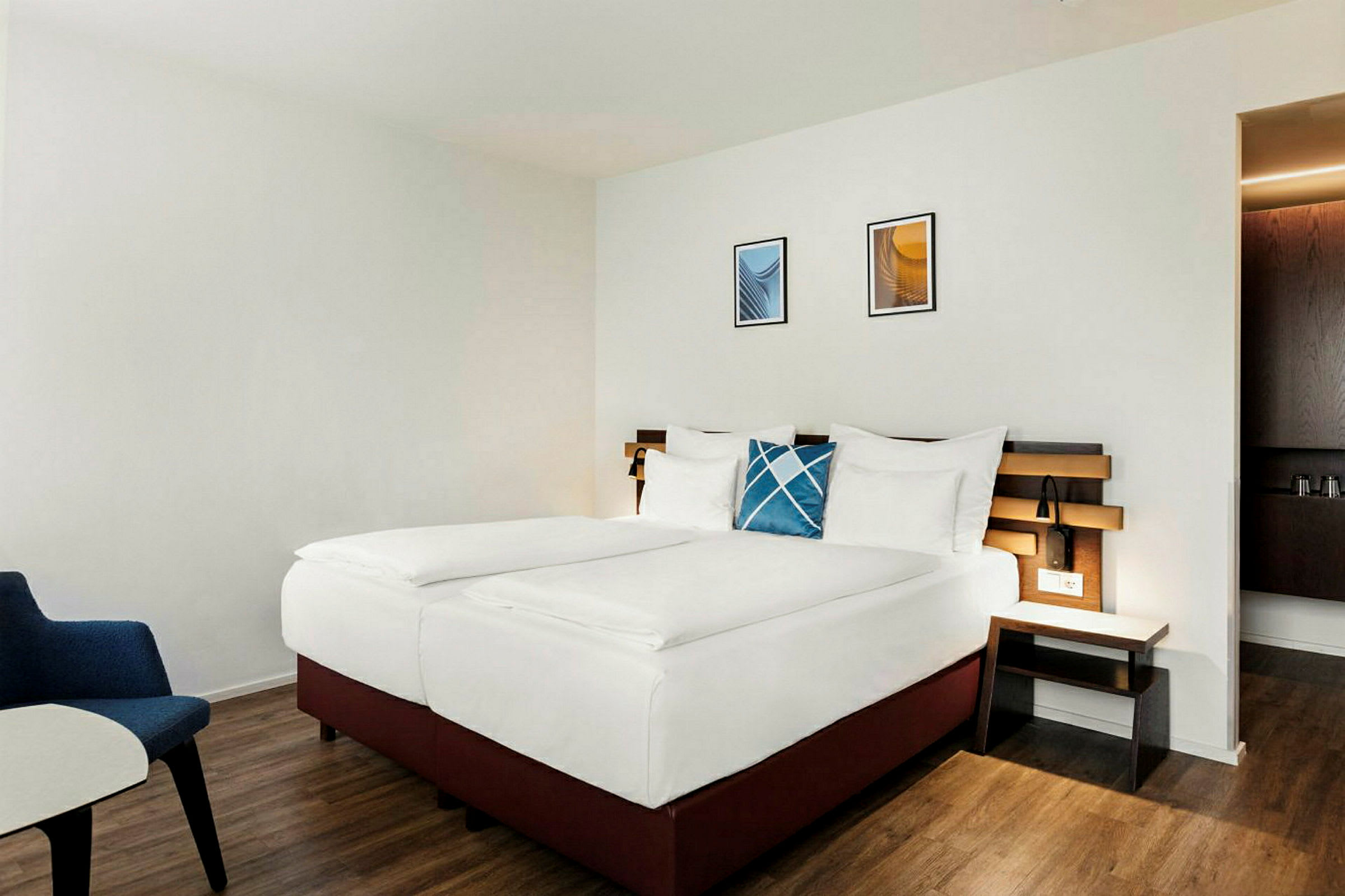 Hotellihuone, jossa on parivuode, valkoiset vuodevaatteet ja sininen koristeellinen tyyny. Sängynpääty on puupaneelia ja huoneessa on puulattia. (Tekoälyn luoma teksti)
