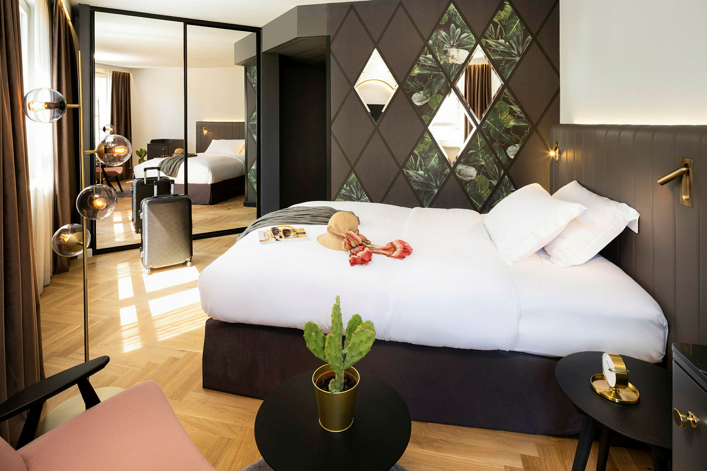Hotellihuone, jossa on suuri sänky, seinäkoristeita ja peili, joka heijastaa huonetta. Matkalaukku seisoo vieressä, ja pöydällä on pieni kaktus. (Tekoälyn luoma teksti)