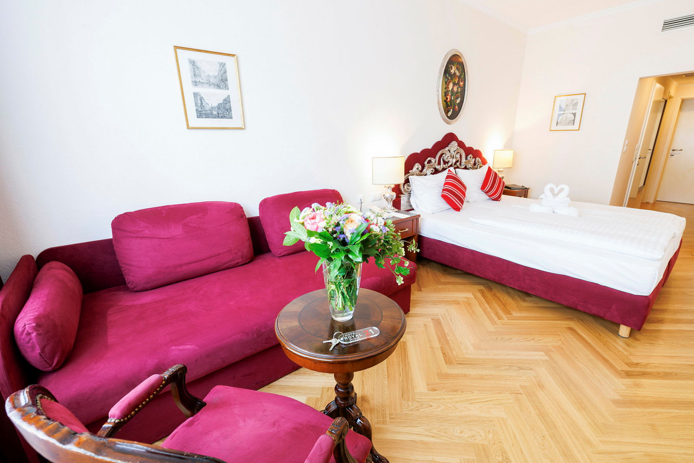 Hotellihuone, jossa on punainen sohva, pyöreä pöytä kukkakimpulla ja punaisin yksityiskohdin varustettu sänky. (Tekoälyn luoma teksti)