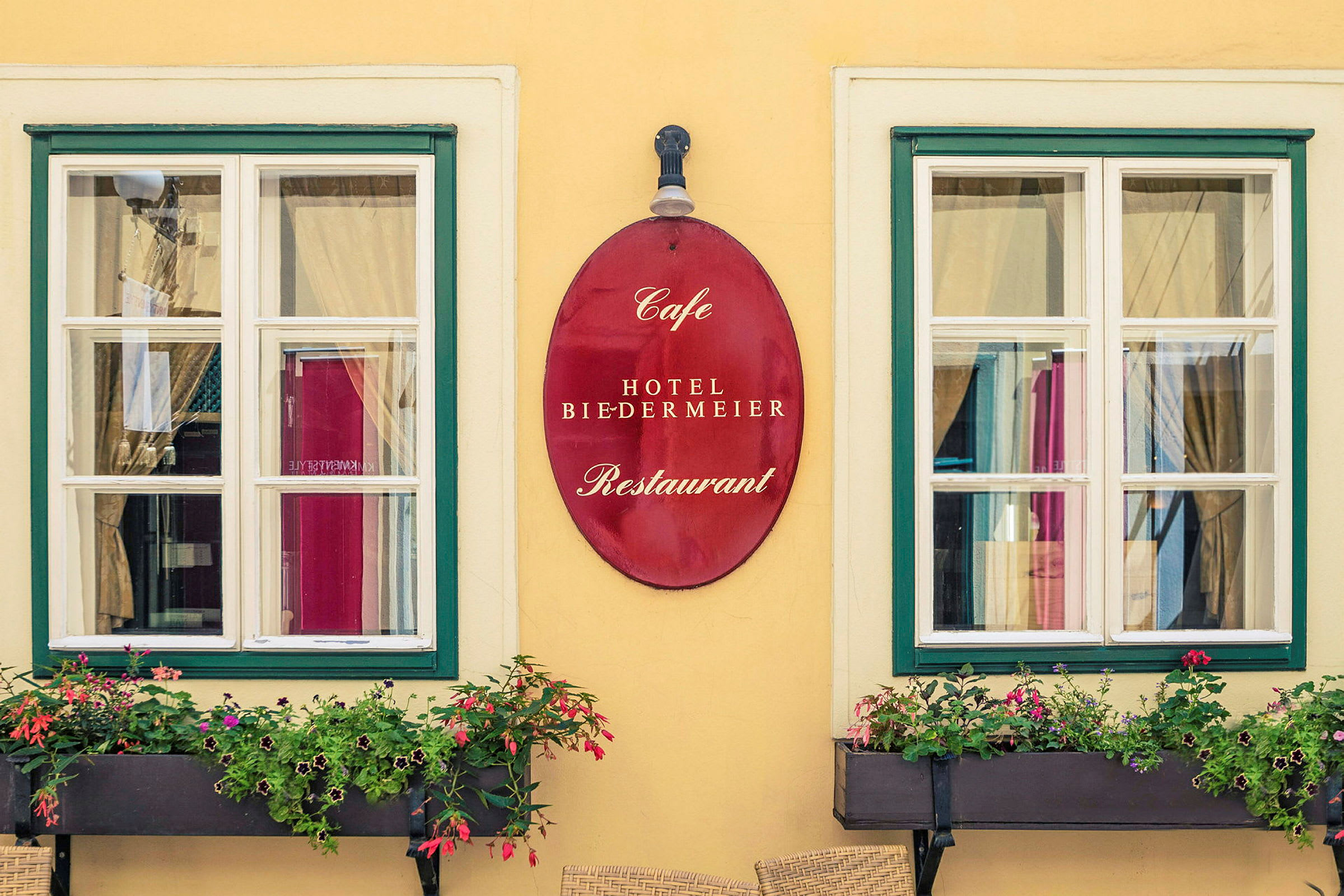 Kaksi ikkunaa, joissa on valkoiset karmit ja vihreät kehykset, niiden välissä roikkuu punainen kyltti, jossa lukee "Cafe Hotel Biedermeier Restaurant". Ikkunoiden edessä on kukkalaatikoita, joissa on kukkivia kasveja. (Tekoälyn luoma teksti)
