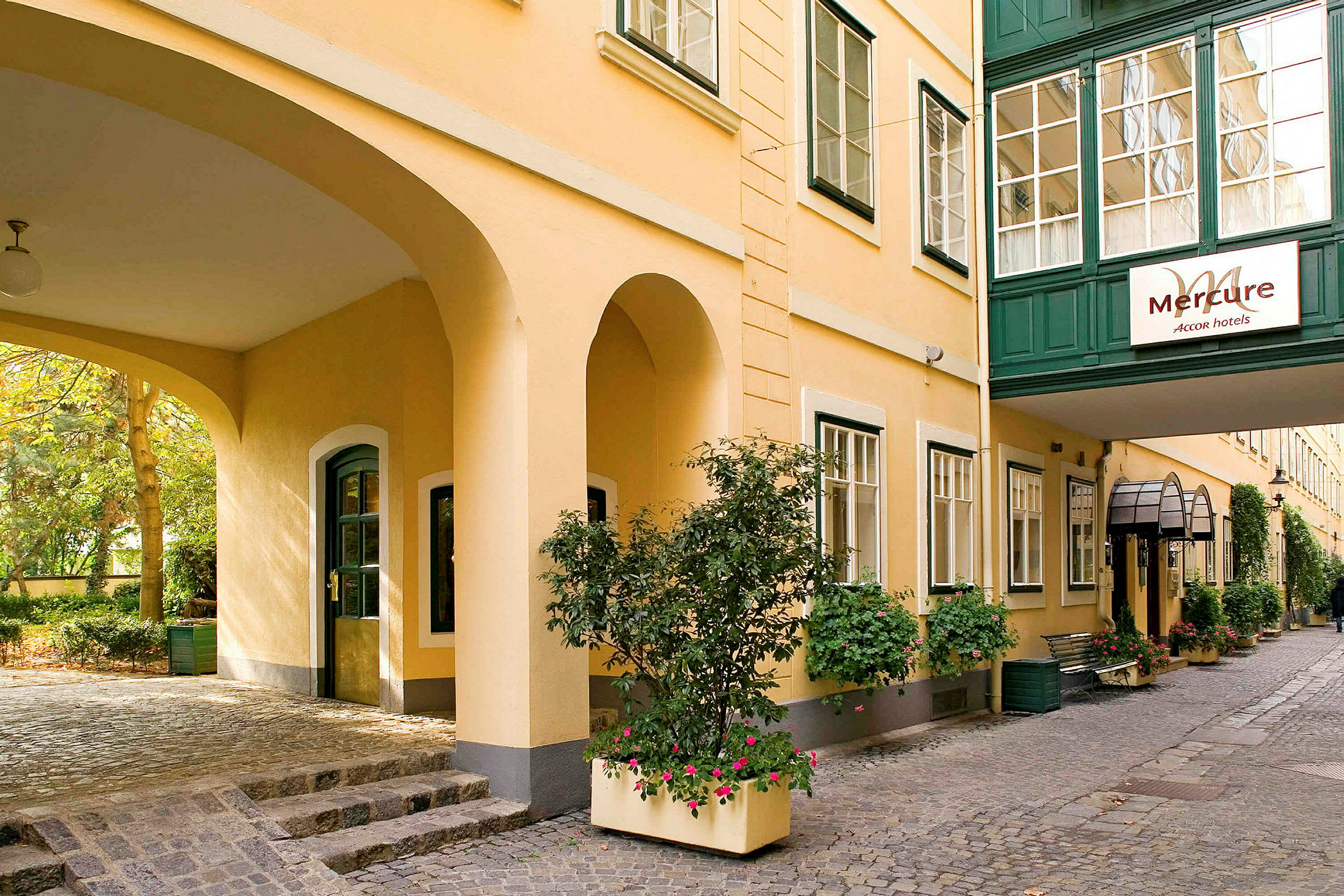Mercure-hotellin sisäpihan näkymä, jossa keltaiset seinät ja vihreät ikkunakehykset, koristeltu kukilla ja vihreällä kasvillisuudella. (Tekoälyn luoma teksti)