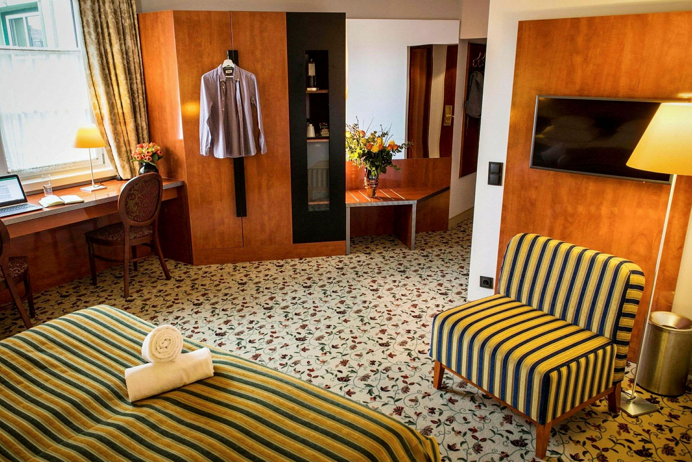 Hotellihuone, jossa on kirjoituspöytä ikkunan vieressä, tuoli, TV-kaappi, nojatuoli ja raidallinen vuode. Seinällä roikkuu paita. (Tekoälyn luoma teksti)