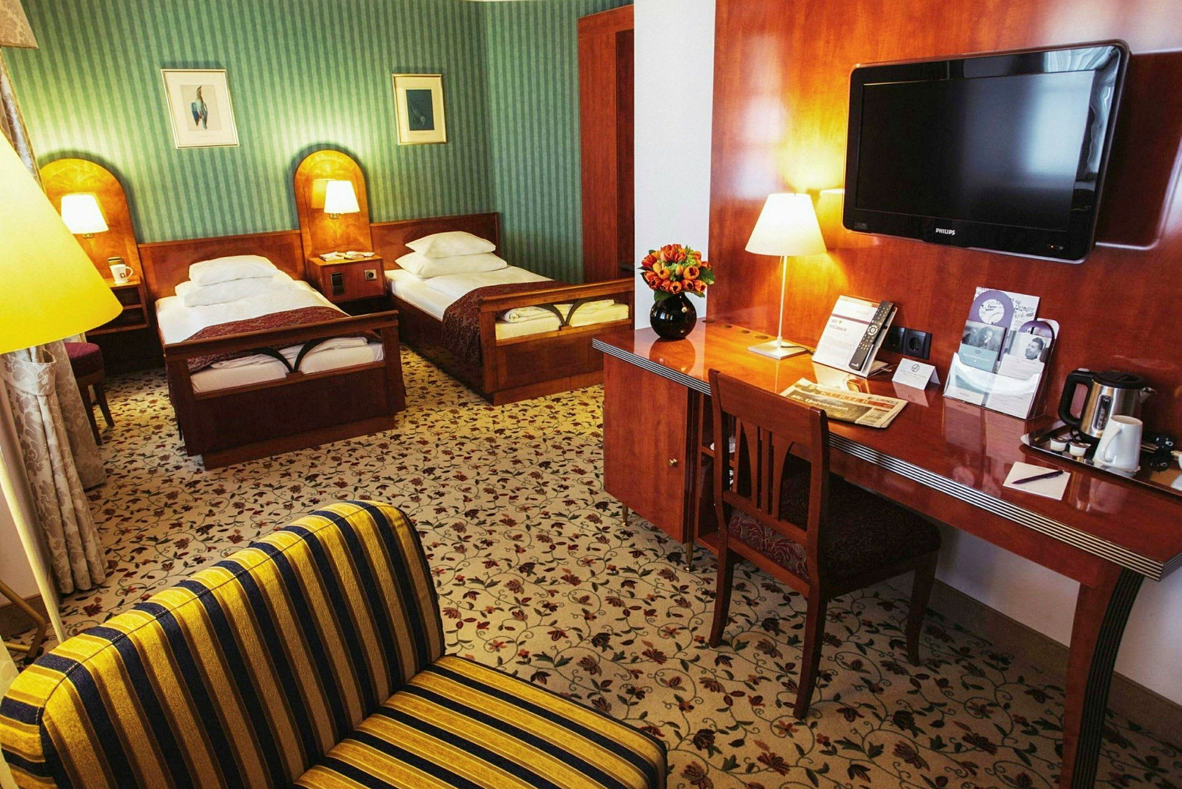 Hotellihuone, jossa on kaksi yhden hengen vuodetta, työpöytä kukilla ja televisio. (Tekoälyn luoma teksti)