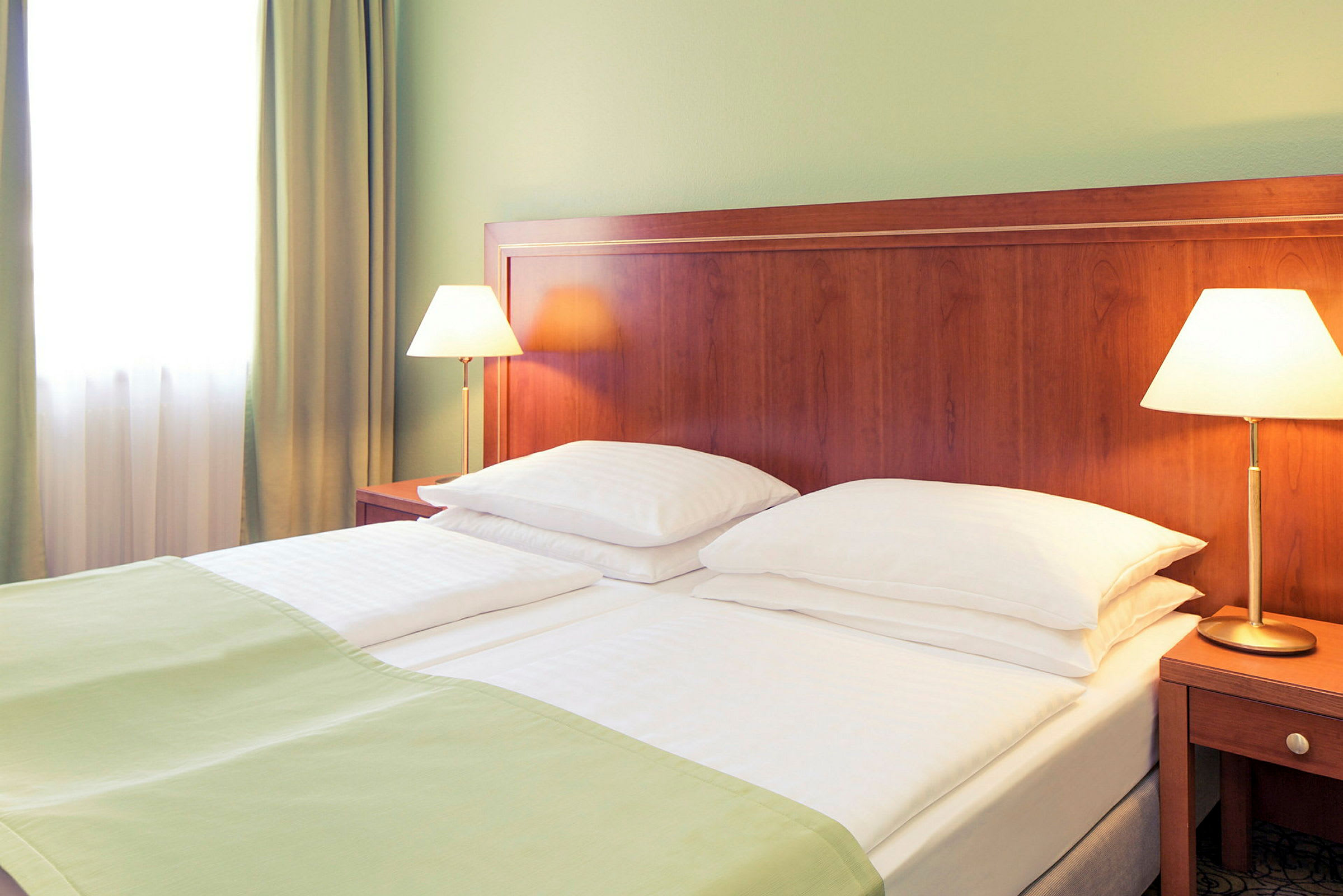 Hotellihuone, jossa on parivuode, valkoiset vuodevaatteet ja vihreät peitot. Kaksi sänkylamppua seisoo puusta tehdyillä yöpöydillä vuoteen vieressä. (Tekoälyn luoma teksti)
