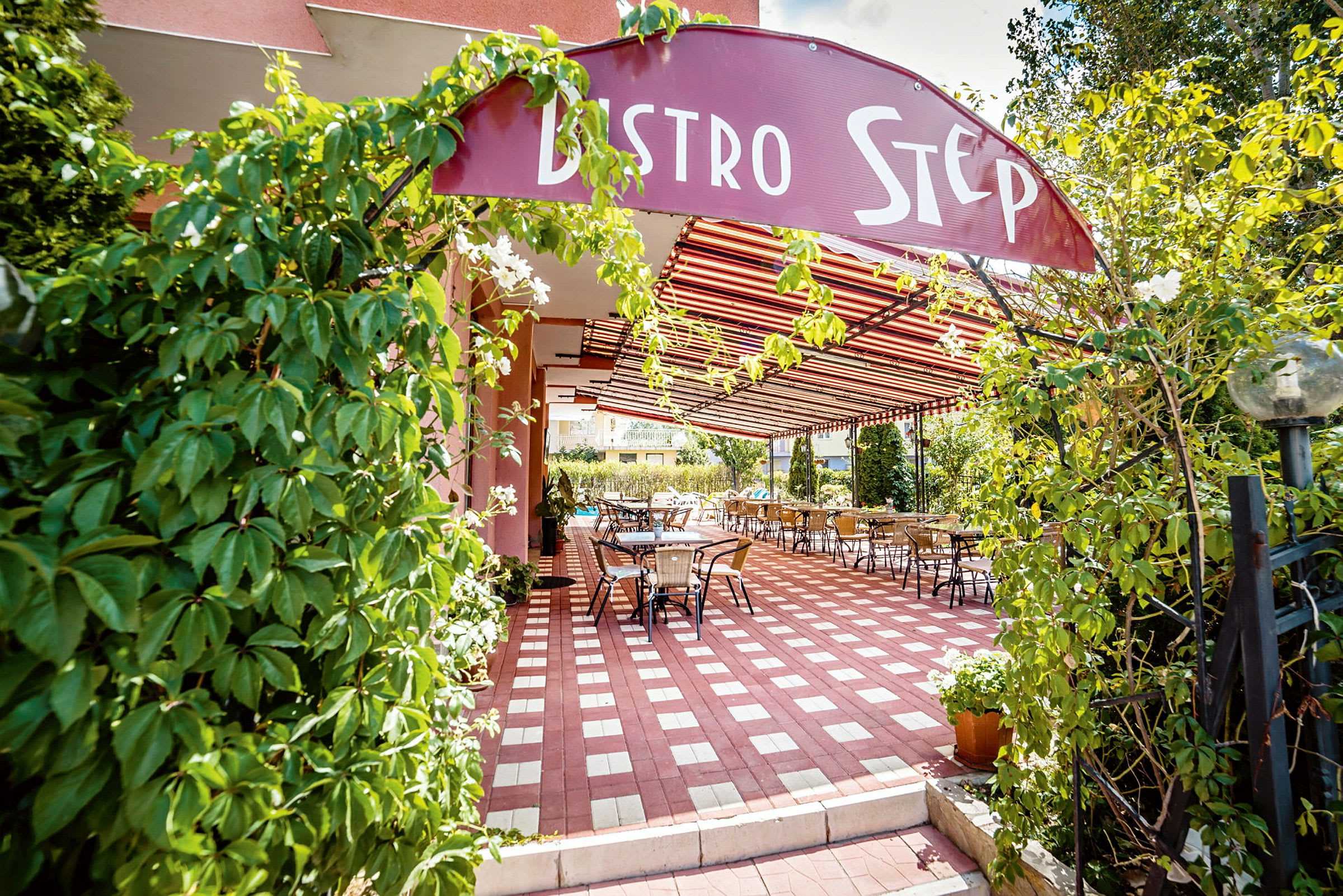 Ravintolan sisäänkäynti, jossa kyltti "Bistro Step", ympärillä vihreää kasvillisuutta ja ulkoterassi katoksen alla. (Tekoälyn luoma teksti)