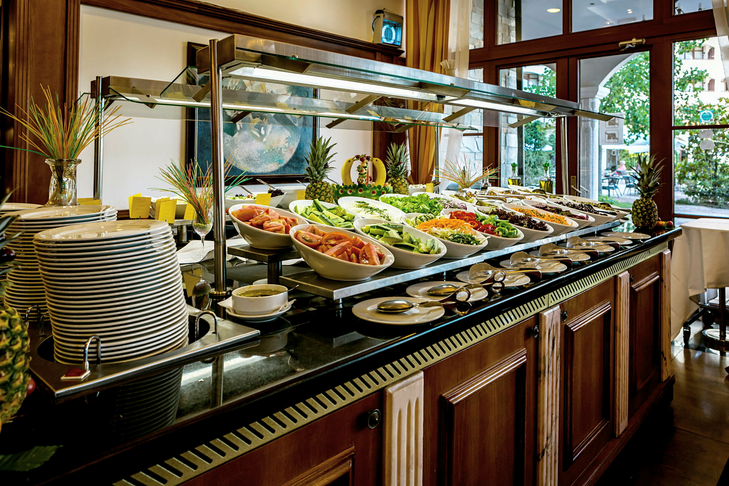 Buffet, jossa on vihanneksia, pastaruokia ja lautasia. Taustalla näkyy ikkunasta tulevaa valoa. (Tekoälyn luoma teksti)