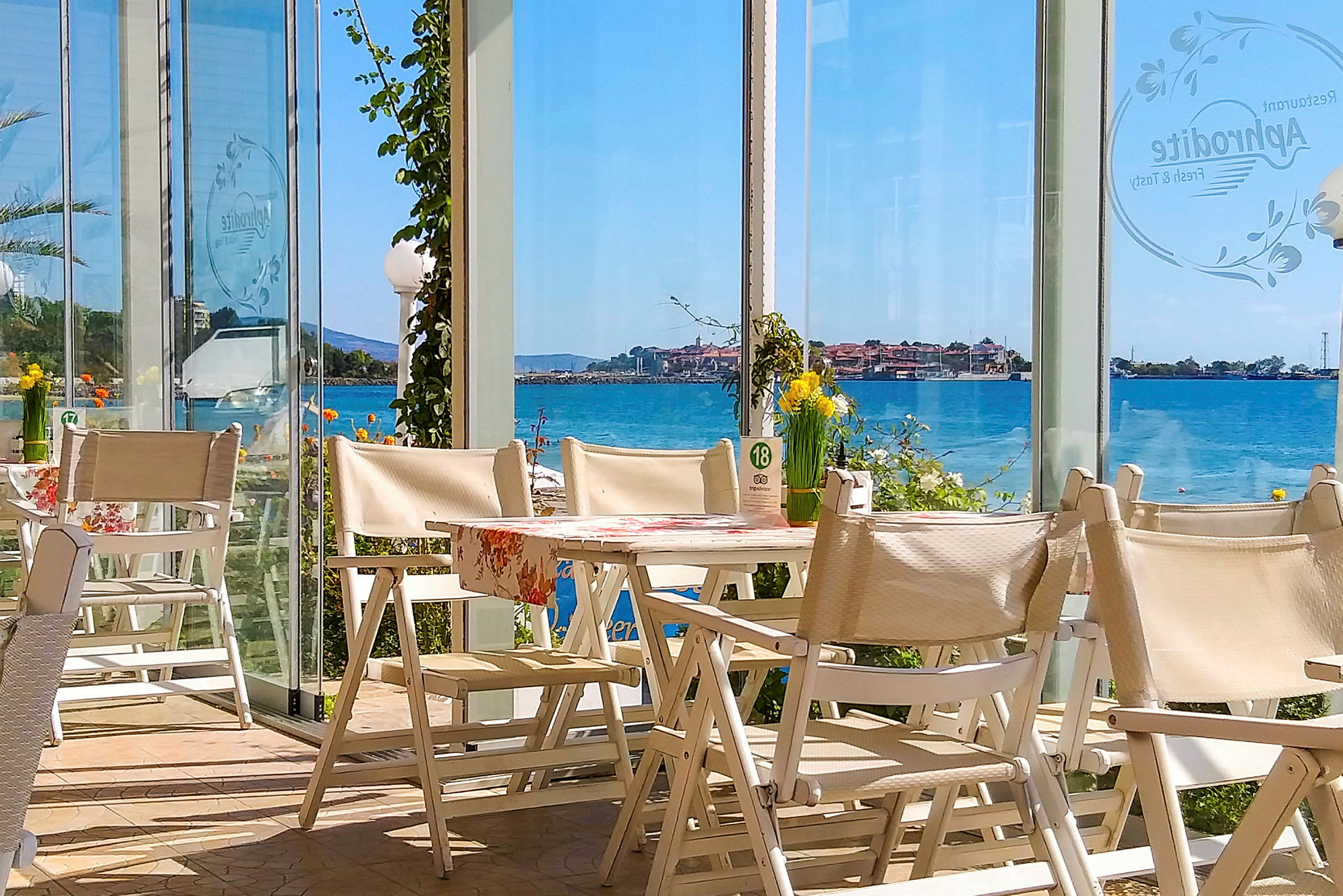 Näköala ravintolasta, jossa on valkoisia tuoleja ja pöytiä sekä lasiseinät merelle ja rannikkokaupunkiin. (Tekoälyn luoma teksti)