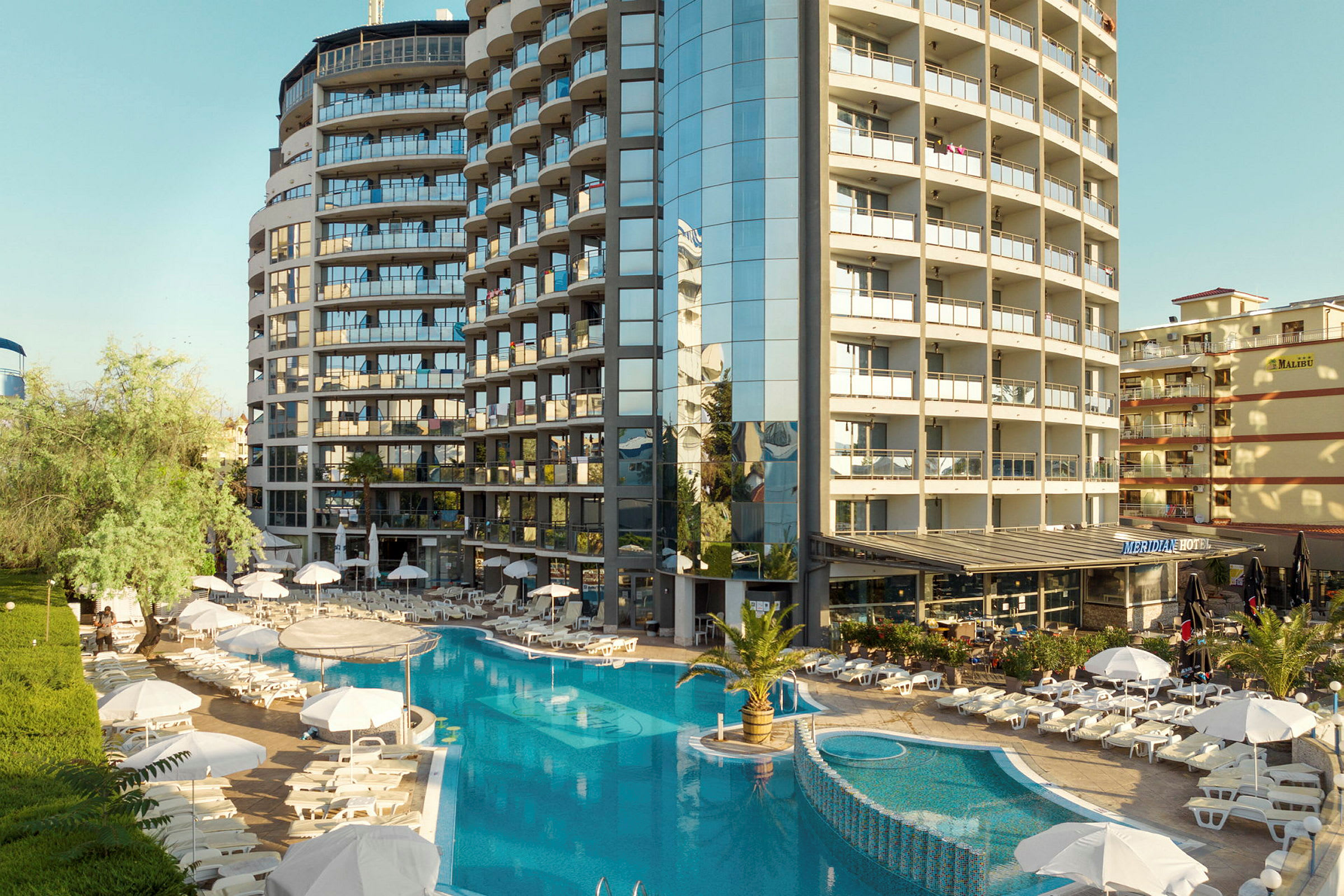 Hotellirakennus, jonka ympärillä on uima-allas ja aurinkotuoleja varjoineen. (Tekoälyn luoma teksti)