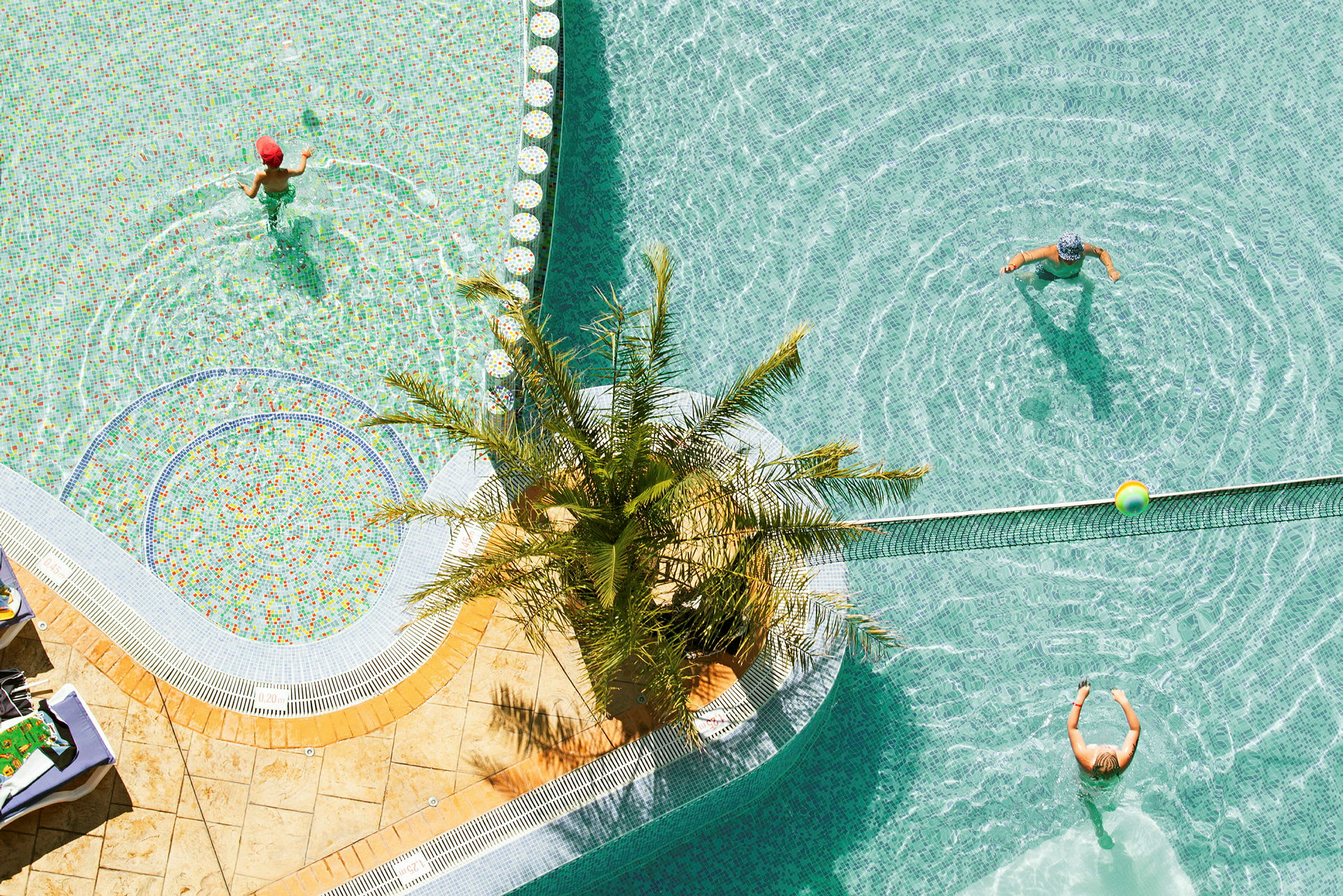 Ulkouima-allas, jossa ihmiset leikkivät vedessä. Palmupuu seisoo altaan reunalla. (Tekoälyn luoma teksti)