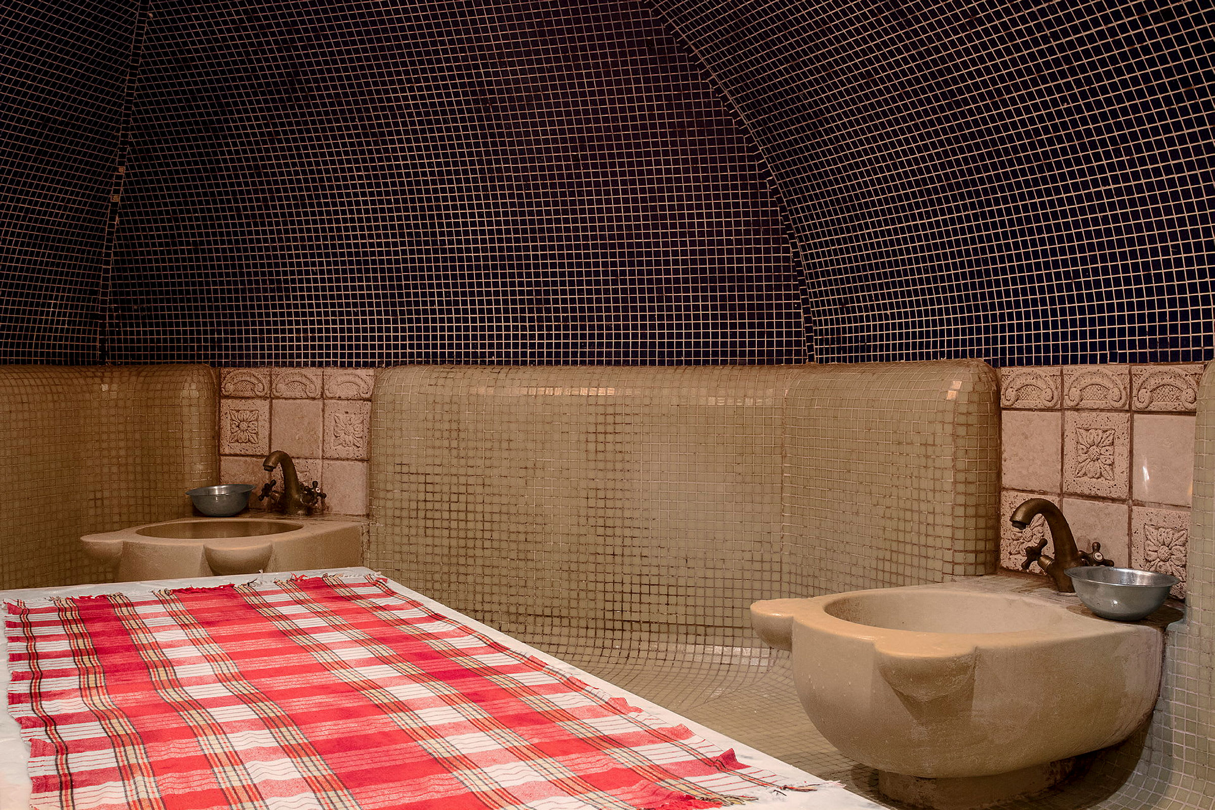 Perinteinen hamam-huone, jossa on kaakeliseinät ja penkki, jonka päällä on ruudullinen pyyhe. (Tekoälyn luoma teksti)