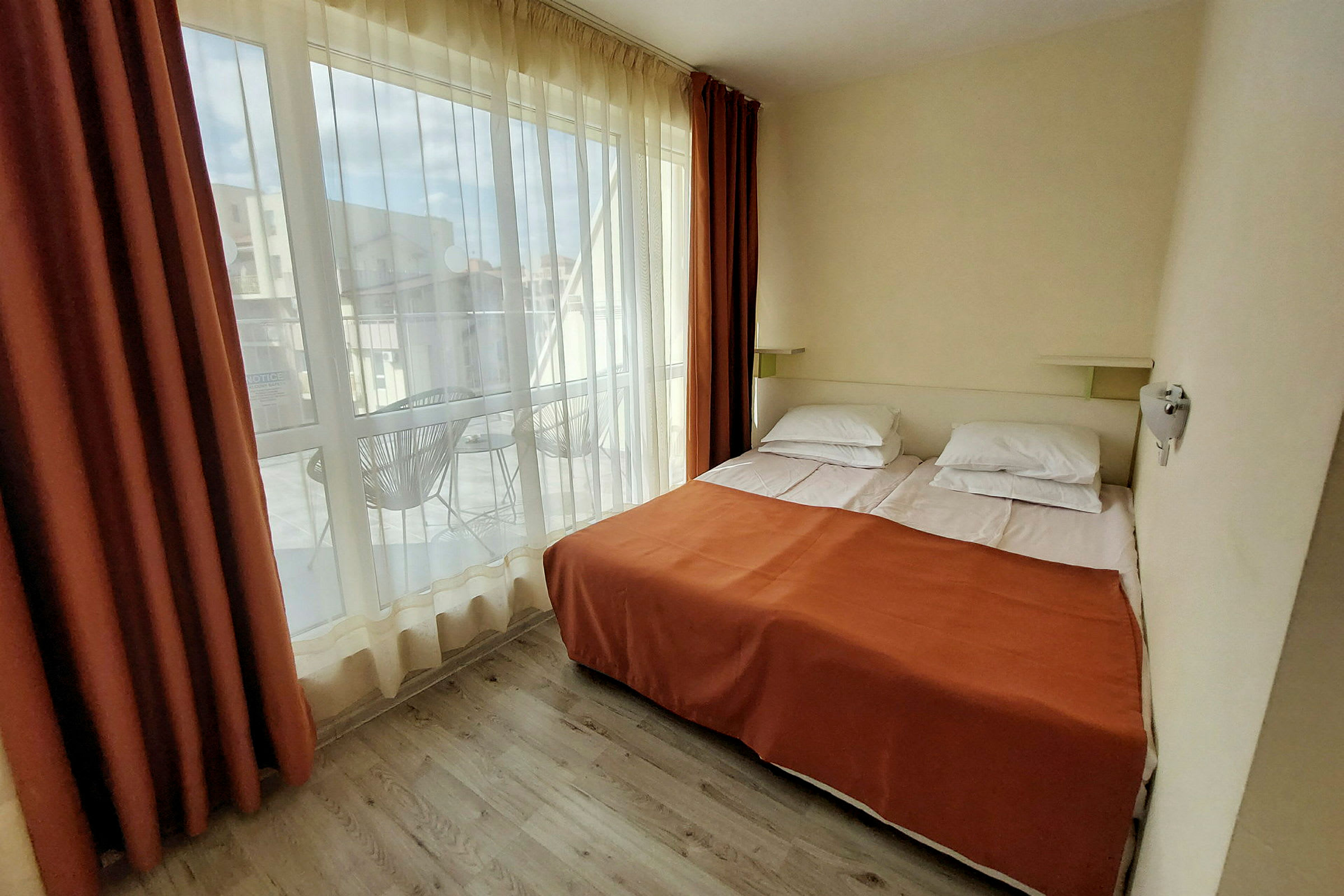 Hotellihuone, jossa kaksi erillistä vuodetta oranssin peitteen alla ja suuret ikkunat, jotka avautuvat parvekkeelle. (Tekoälyn luoma teksti)