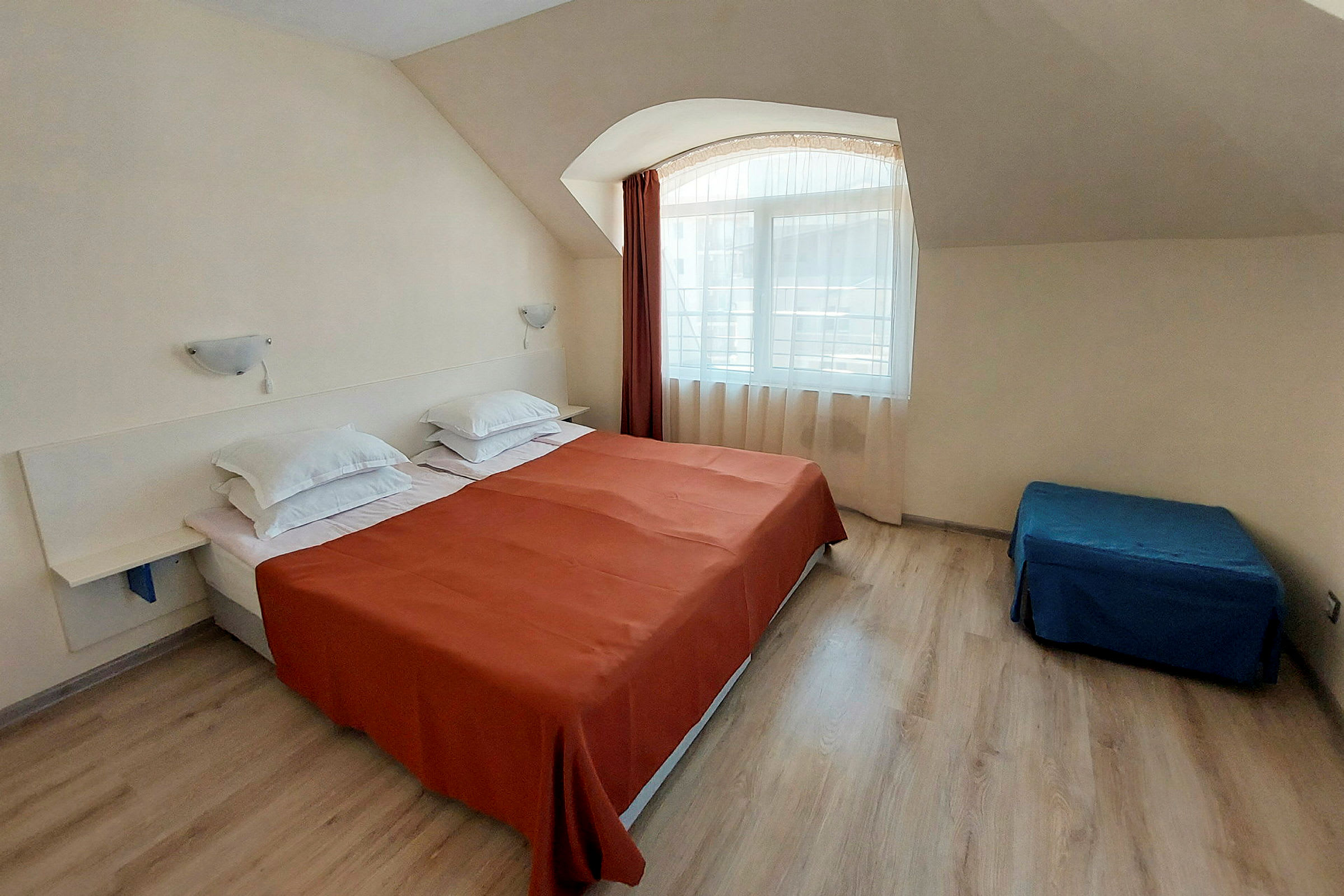 Hotellihuone, jossa on parivuode, jonka päällä on oranssi peitto ja valkoiset tyynyt, sininen istuinrahi ja ikkuna vaaleilla verhoilla. (Tekoälyn luoma teksti)