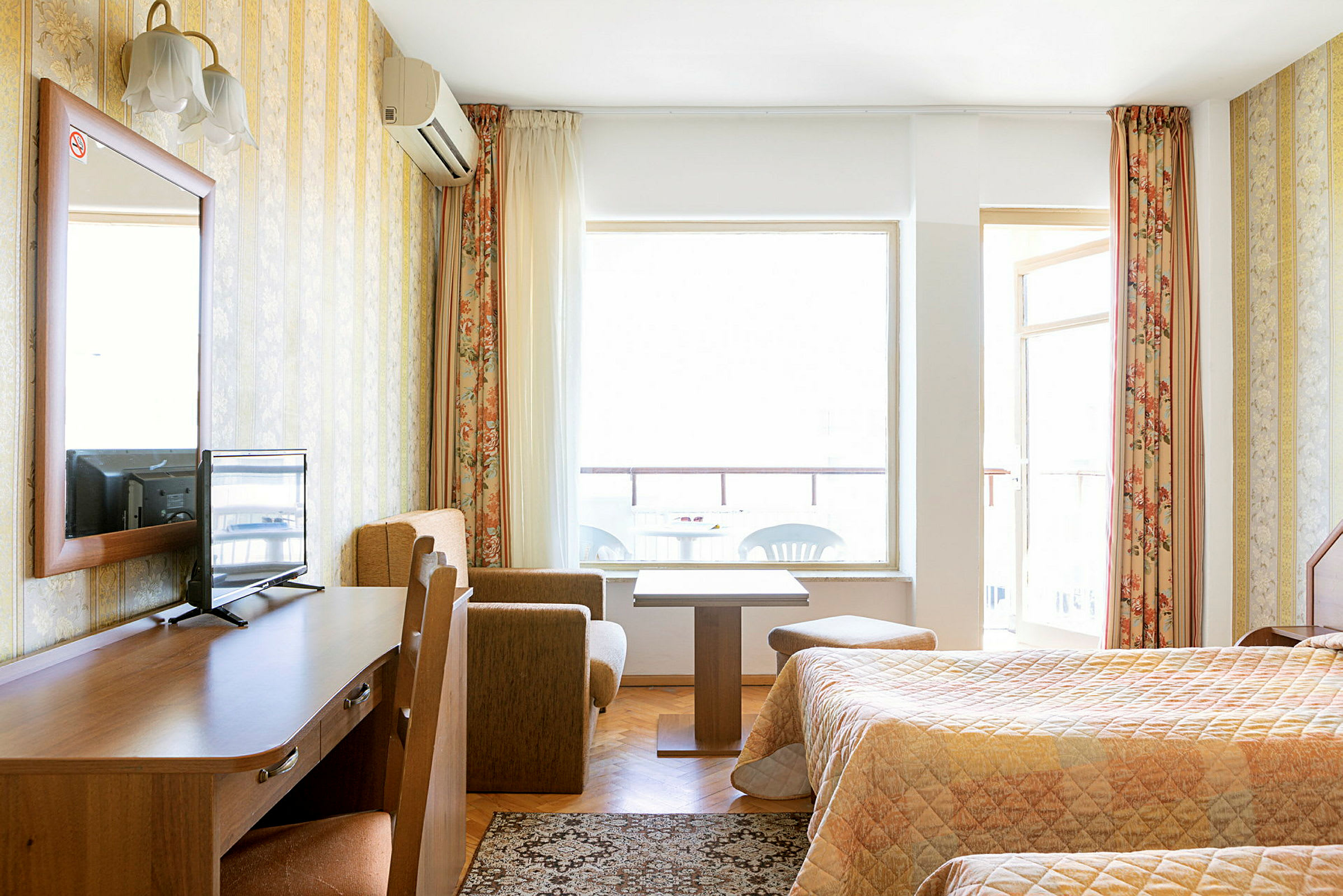 Hotellihuone, jossa on kaksi sänkyä, puinen työpöytä, jolla on TV, pieni sohvanurkkaus sohvapöydällä ja parveke taustalla. (Tekoälyn luoma teksti)