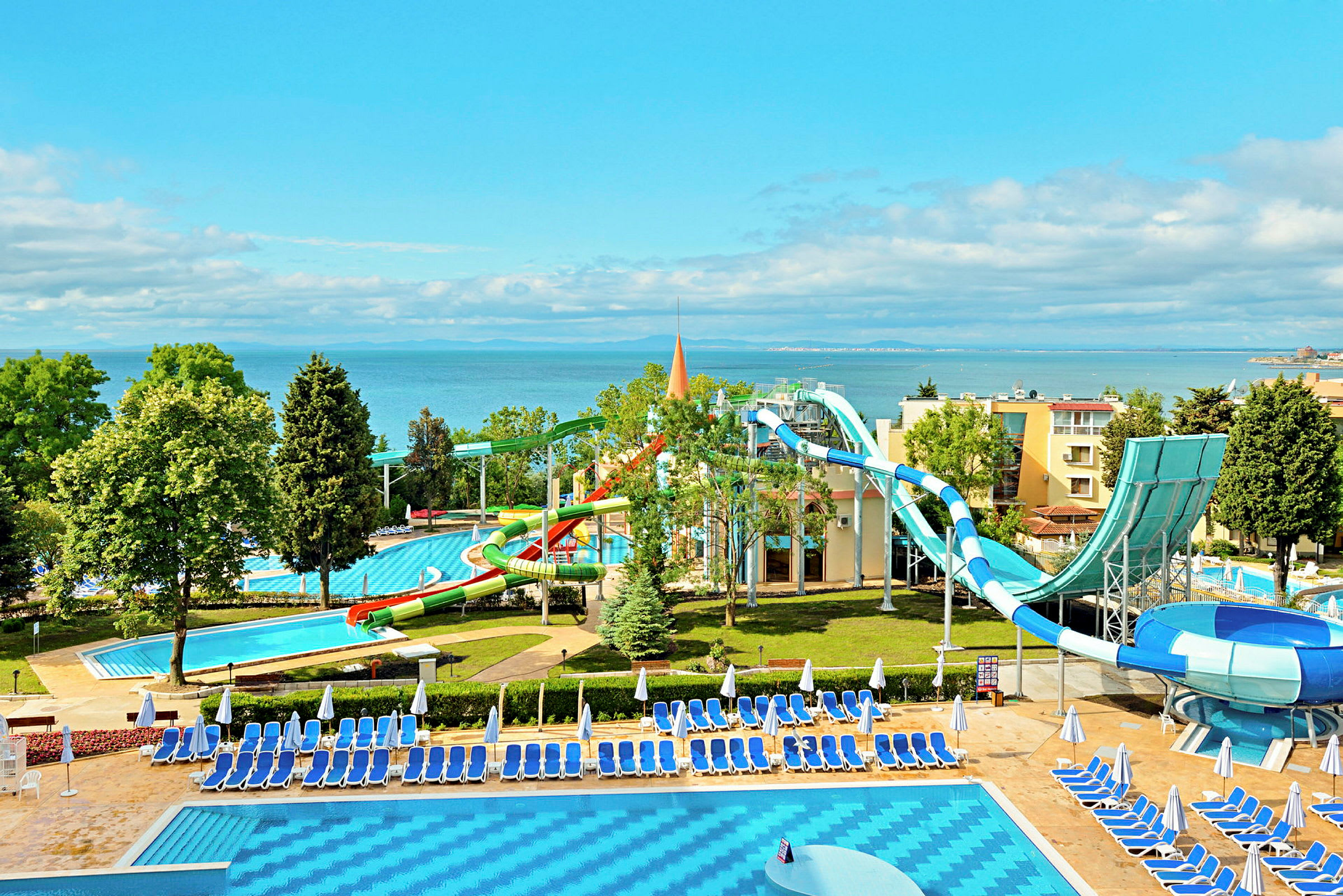 Vesipuisto meren rannalla, jossa on vesiliukumäkiä ja uima-altaita ympäröitynä aurinkotuoleilla ja -varjoilla. (Tekoälyn luoma teksti)