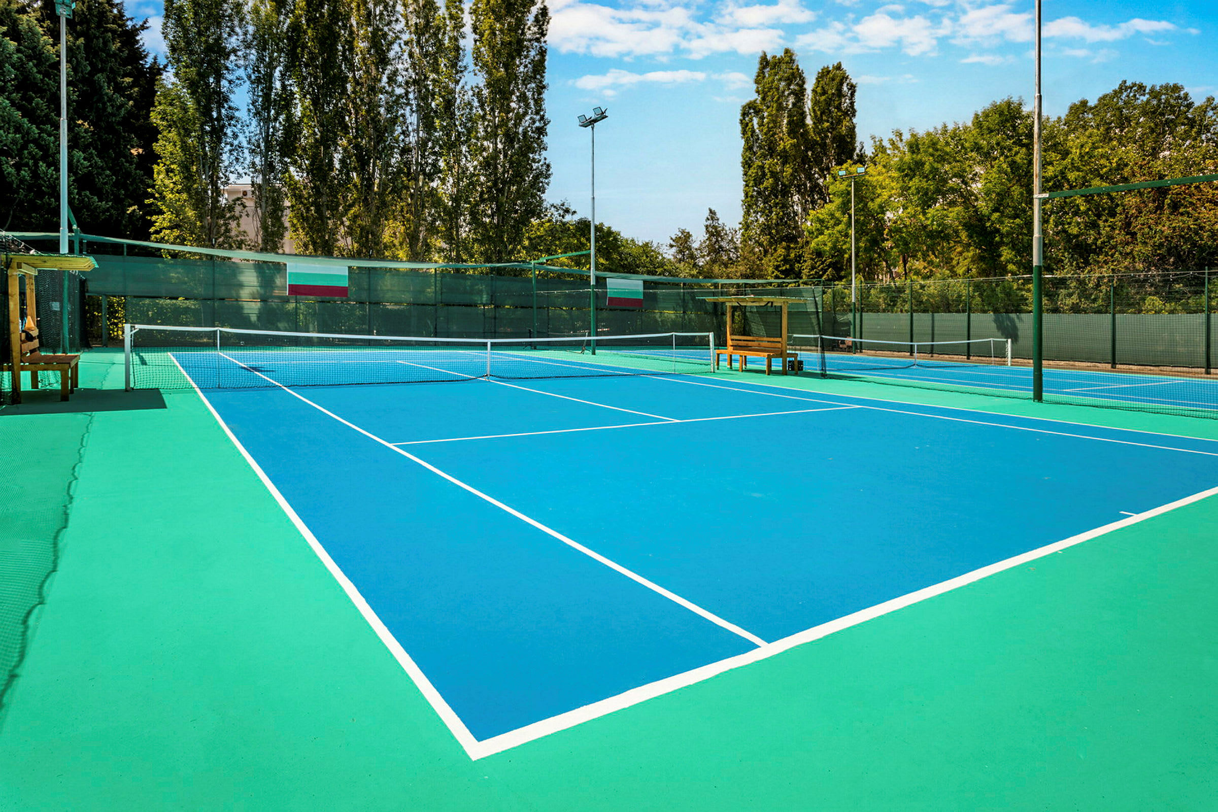 Kuva tenniskentästä, jossa on sininen pinta ja valkoiset linjat, ympäröitynä puilla ja aidalla. (Tekoälyn luoma teksti)