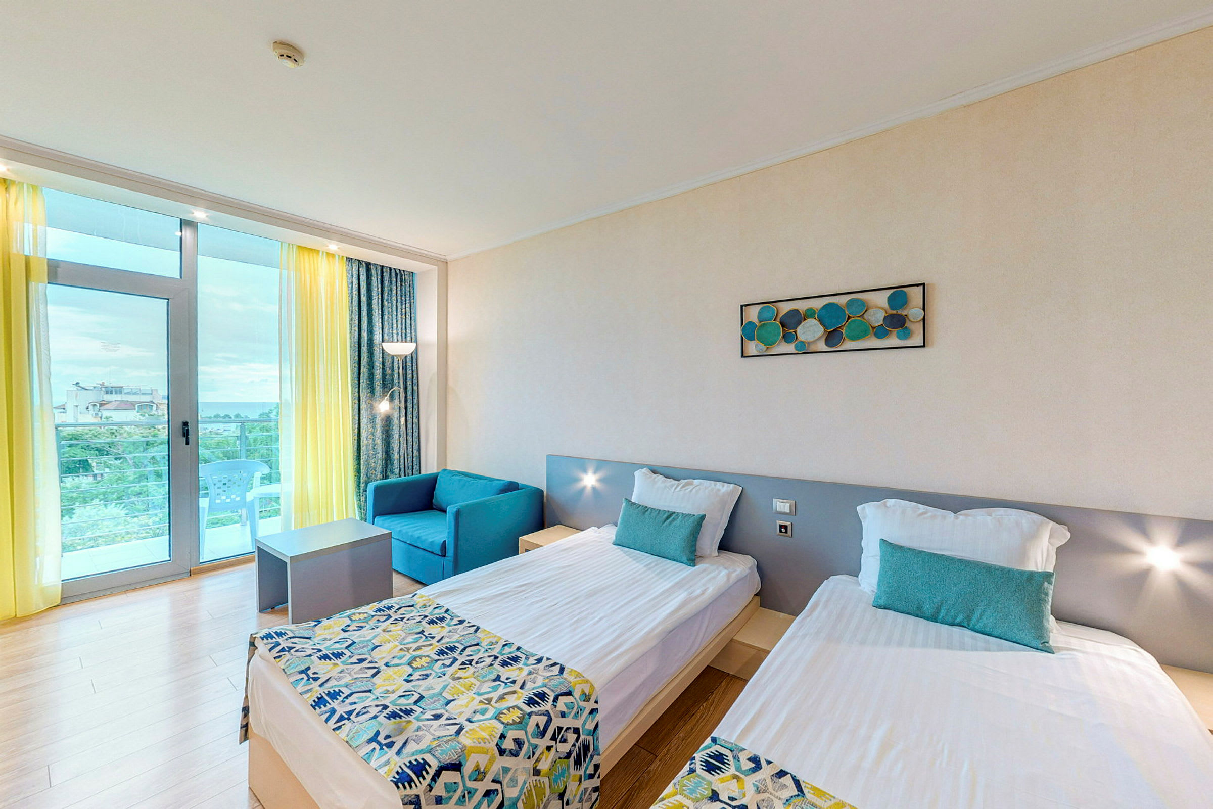 Hotellihuone, jossa on kaksi yhden hengen sänkyä, sininen nojatuoli ja pöytä. Huoneessa on vaaleat seinät ja suuri ikkuna, jossa on keltainen verho ja parveke. (Tekoälyn luoma teksti)