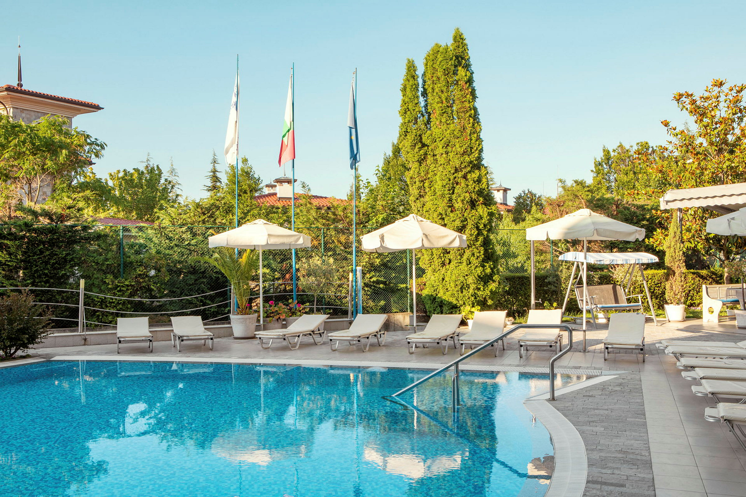 Ulkouima-allas, jossa on aurinkotuoleja ja aurinkovarjoja hotellilla, ympärillä vihreitä puita ja lipputankoja. (Tekoälyn luoma teksti)