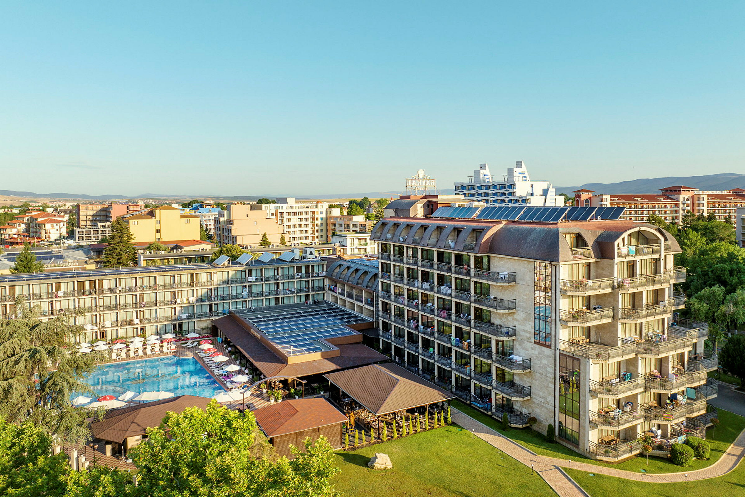 Ilmakuva hotellista, jossa on uima-allas ja aurinkotuoleja, ympäröitynä rakennuksilla ja vehreydellä. (Tekoälyn luoma teksti)