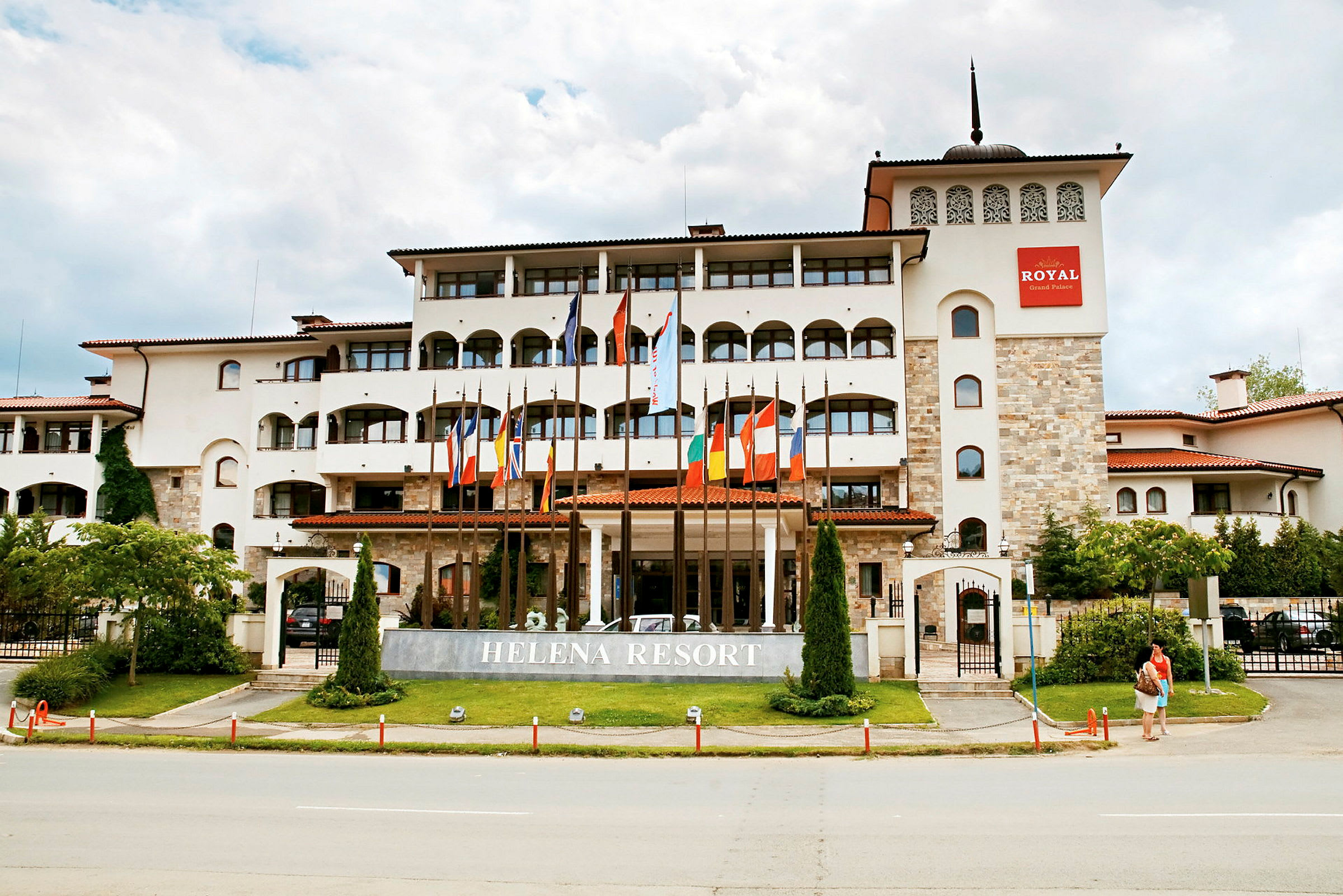 Usean kerroksen moderni hotellirakennus, edessä lippuja ja tekstinä 'Helena Resort' kyltissä. (Tekoälyn luoma teksti)