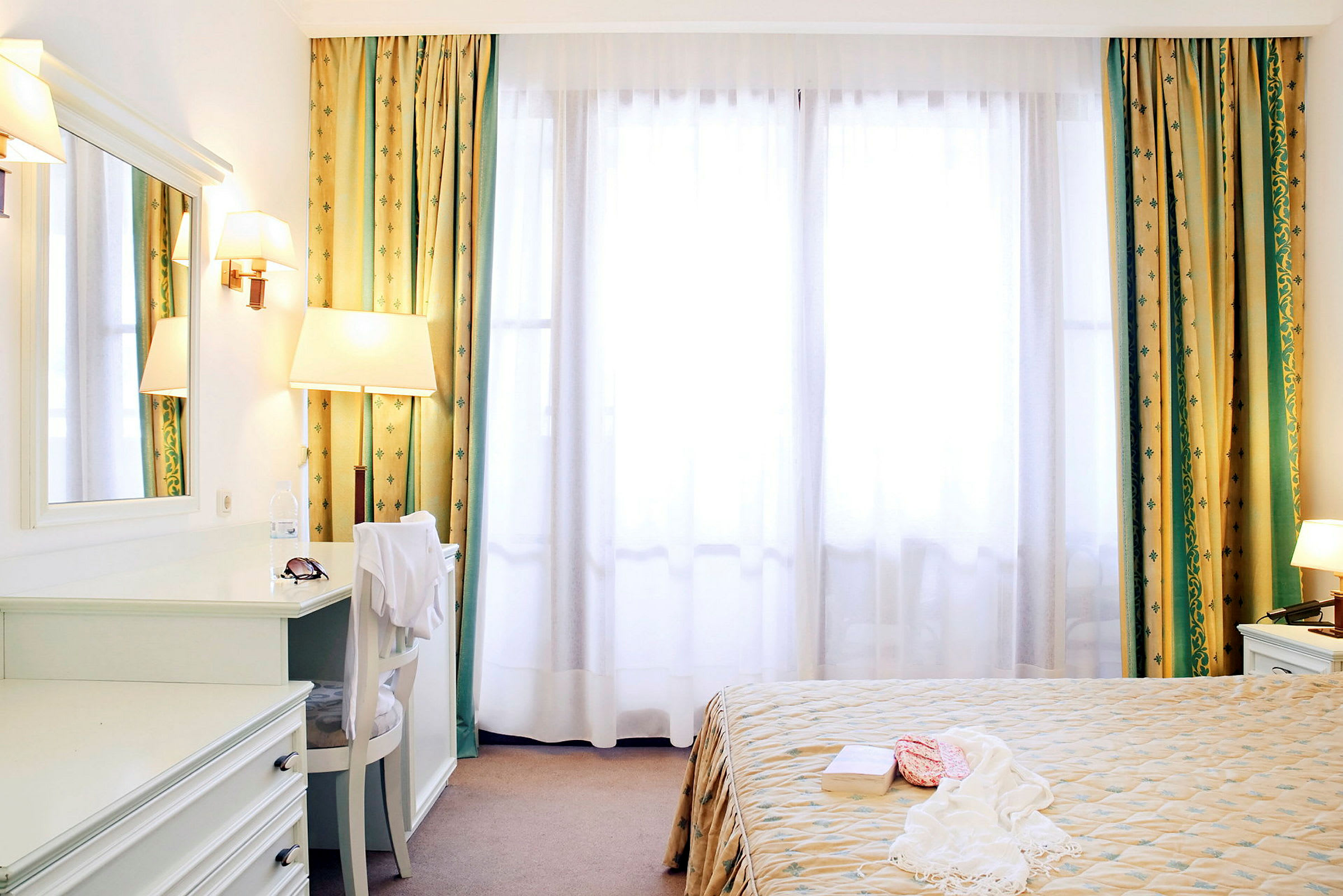 Valoisa hotellihuone, jossa on sänky, peili ja suuret ikkunat verhoilla. (Tekoälyn luoma teksti)
