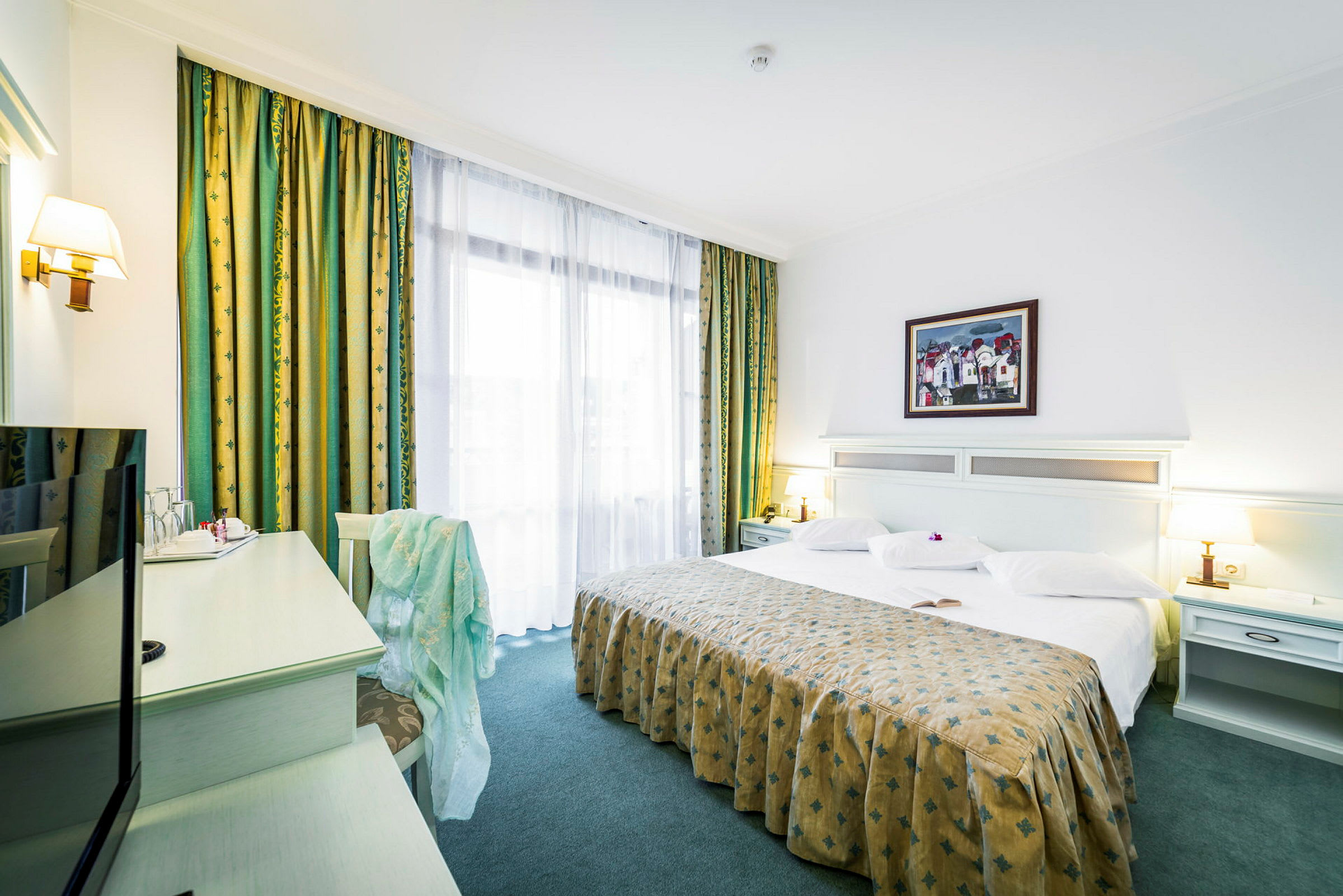 Hotellihuone, jossa on suuri sänky, valkoiset vuodevaatteet ja kuvioitu päiväpeitto. Sängyn vieressä on pöytävalaisimia sekä nurkassa kirjoituspöytä ja tuoli. (Tekoälyn luoma teksti)