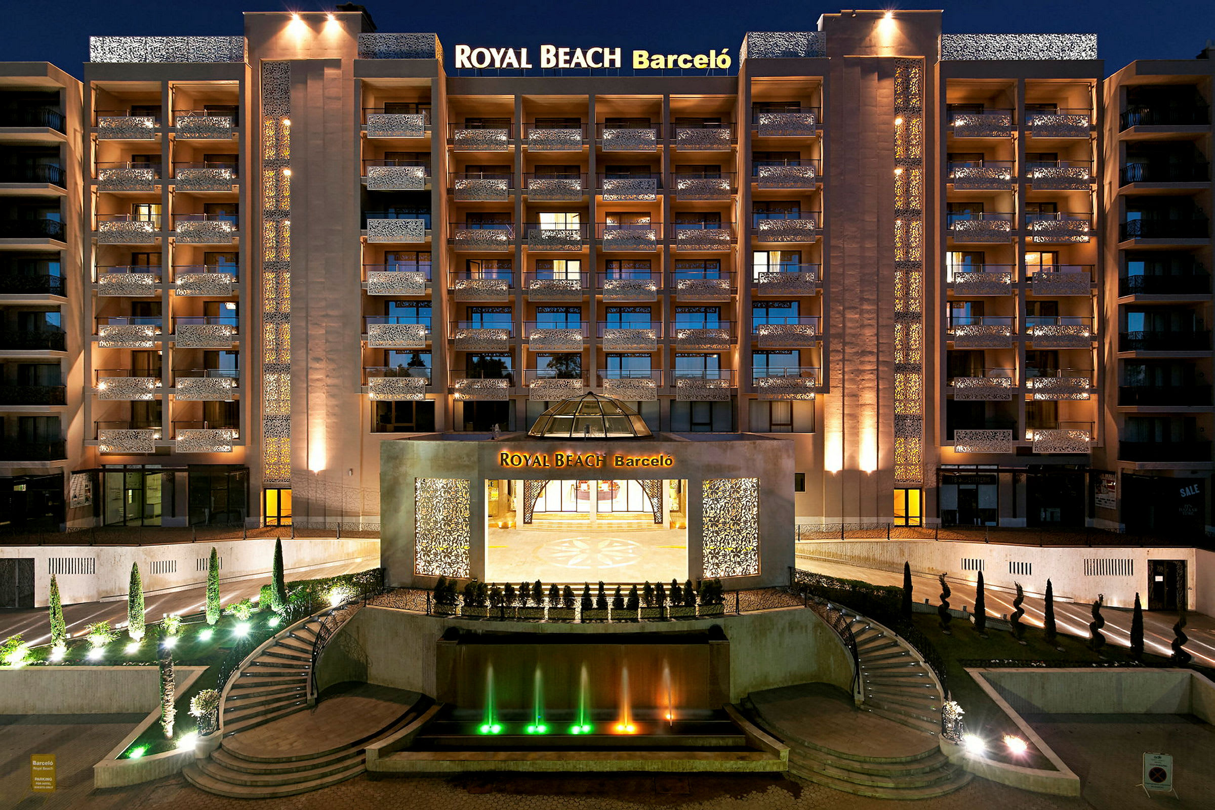 Yönäkymä valaistusta hotellista nimeltä Royal Beach Barceló, etualalla suihkulähde ja portaat. (Tekoälyn luoma teksti)