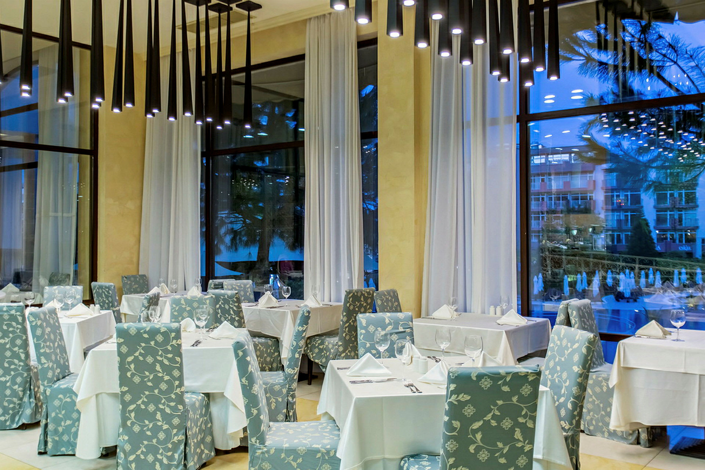 Tyylikäs ravintola, jossa pöydät ovat valkoisilla liinoilla peitettyjä ja tuolit kukallista kuviota, korkeat ikkunat valkoisilla verhoilla. (Tekoälyn luoma teksti)