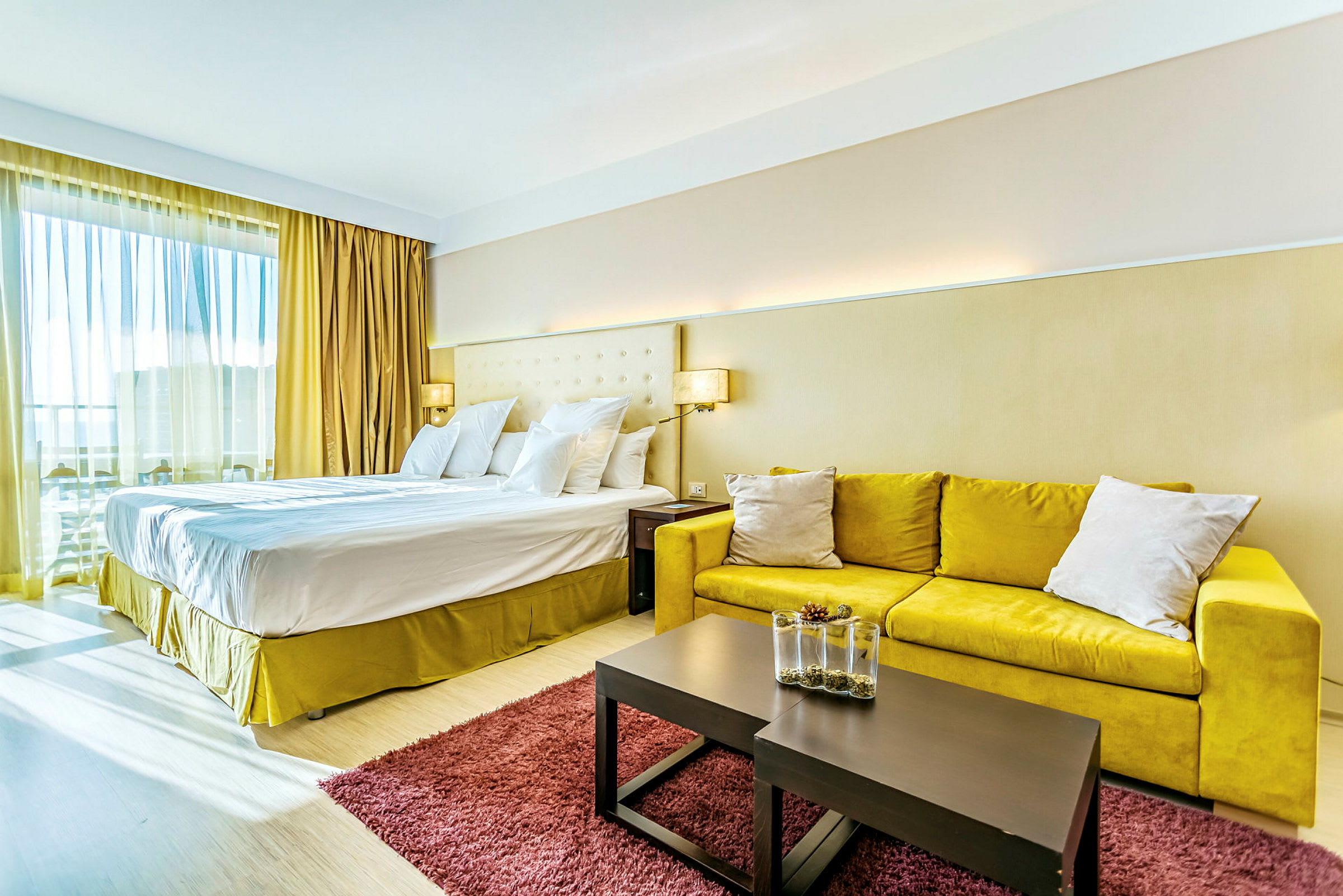 Hotellihuone, jossa suuri parisänky, keltainen sohva, pöytä koristeellisella maljakolla ja punainen matto. (Tekoälyn luoma teksti)