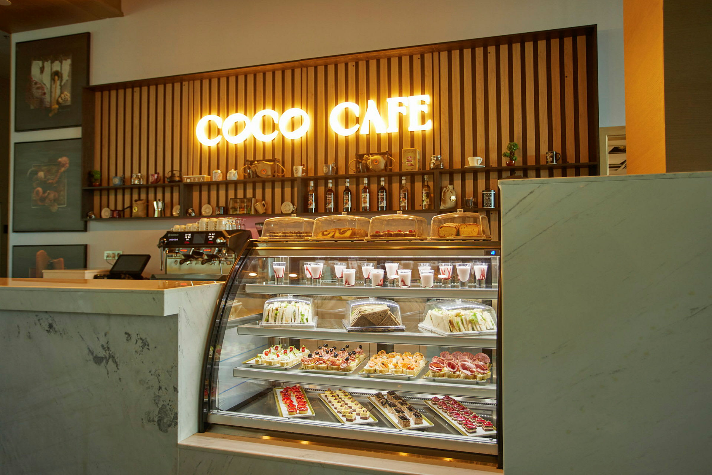 Kahvilan tiski, jossa valaistu kyltti 'Coco Cafe' ja valikoima leivonnaisia ja juomia lasin takana. (Tekoälyn luoma teksti)