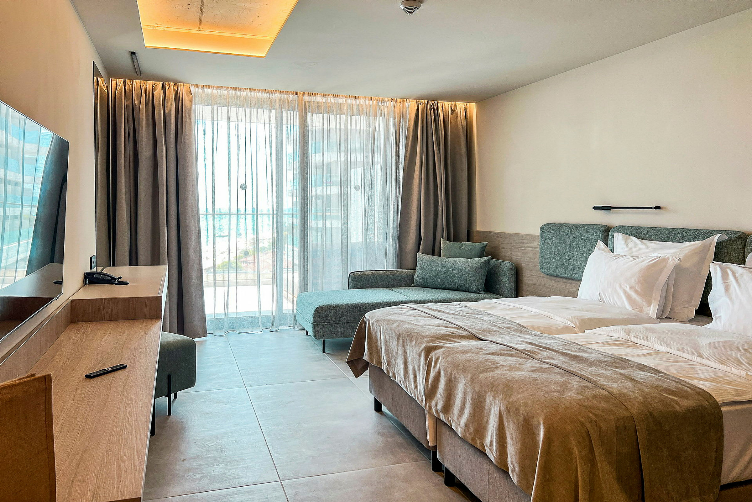 Hotellihuone, jossa on parivuode, sohva ja työpöytä ikkunan edessä vaaleilla verhoilla. (Tekoälyn luoma teksti)