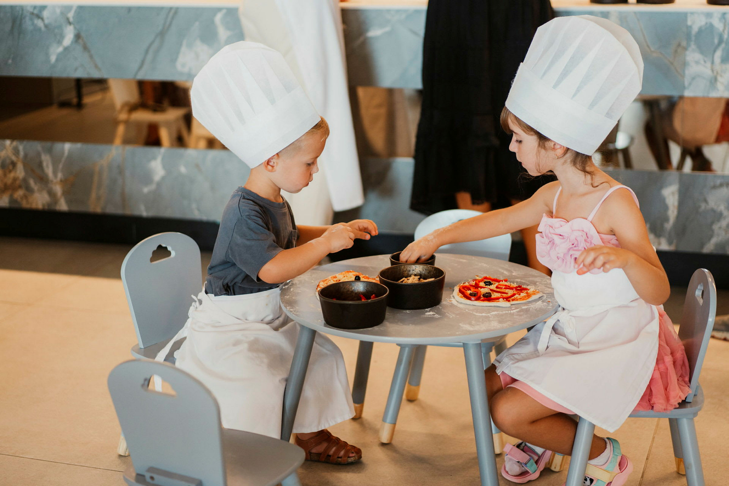 Kaksi lasta kokin hatuilla ja esiliinoilla koristelemassa pizzoja pöydän ääressä. (Tekoälyn luoma teksti)