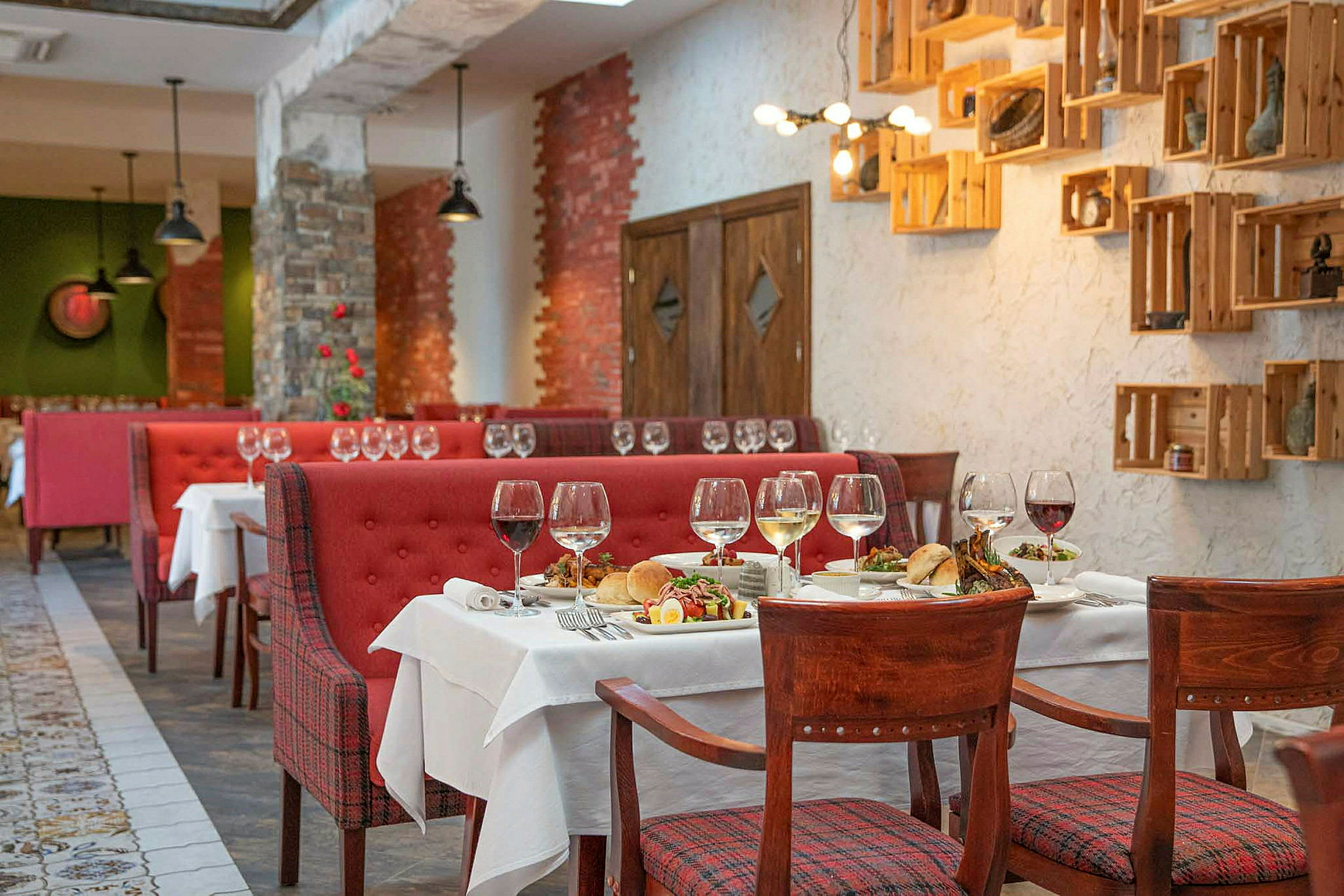 Ravintola, jossa on punaisia sohvia, puutuoleja ja katettuja pöytiä viinilaseilla ja aterioilla. (Tekoälyn luoma teksti)