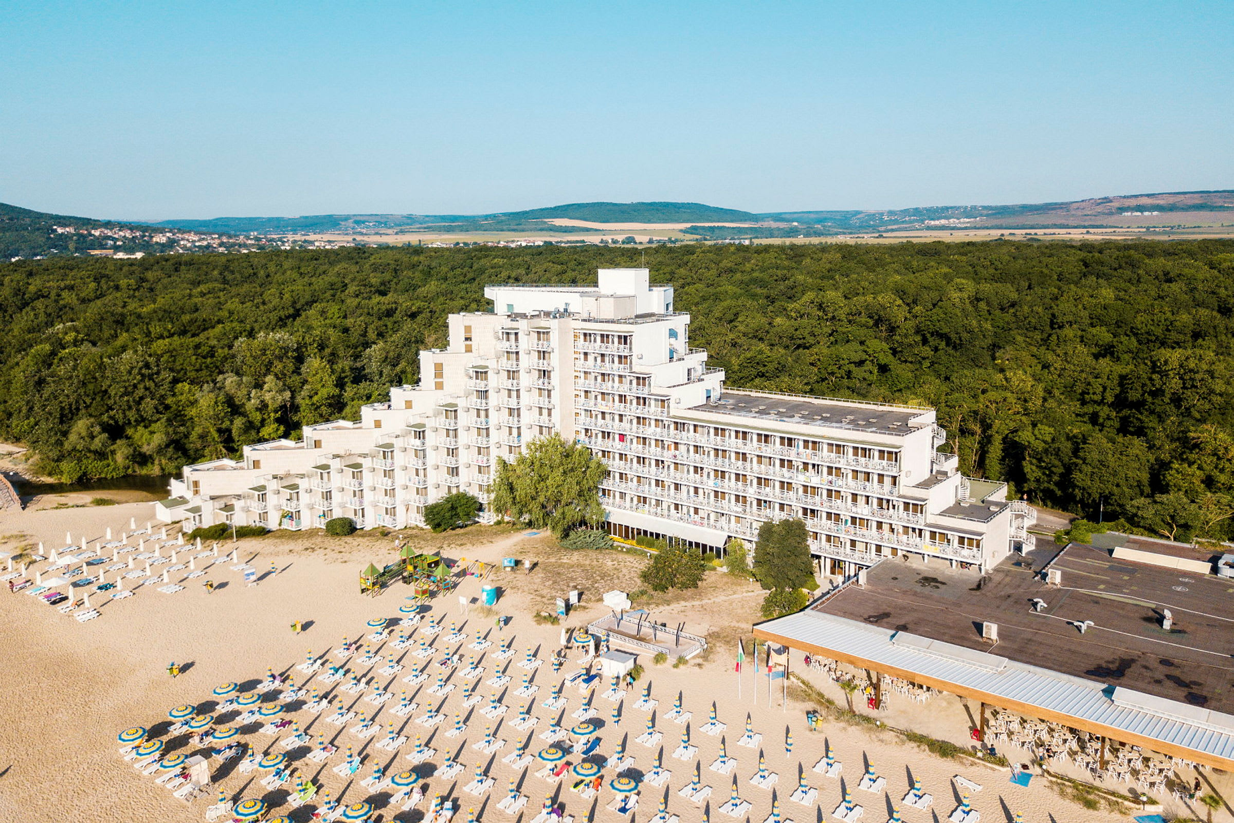Kuva hotellista rannalla, jossa on aurinkotuoleja ja aurinkovarjoja, metsän ympäröimänä. (Tekoälyn luoma teksti)