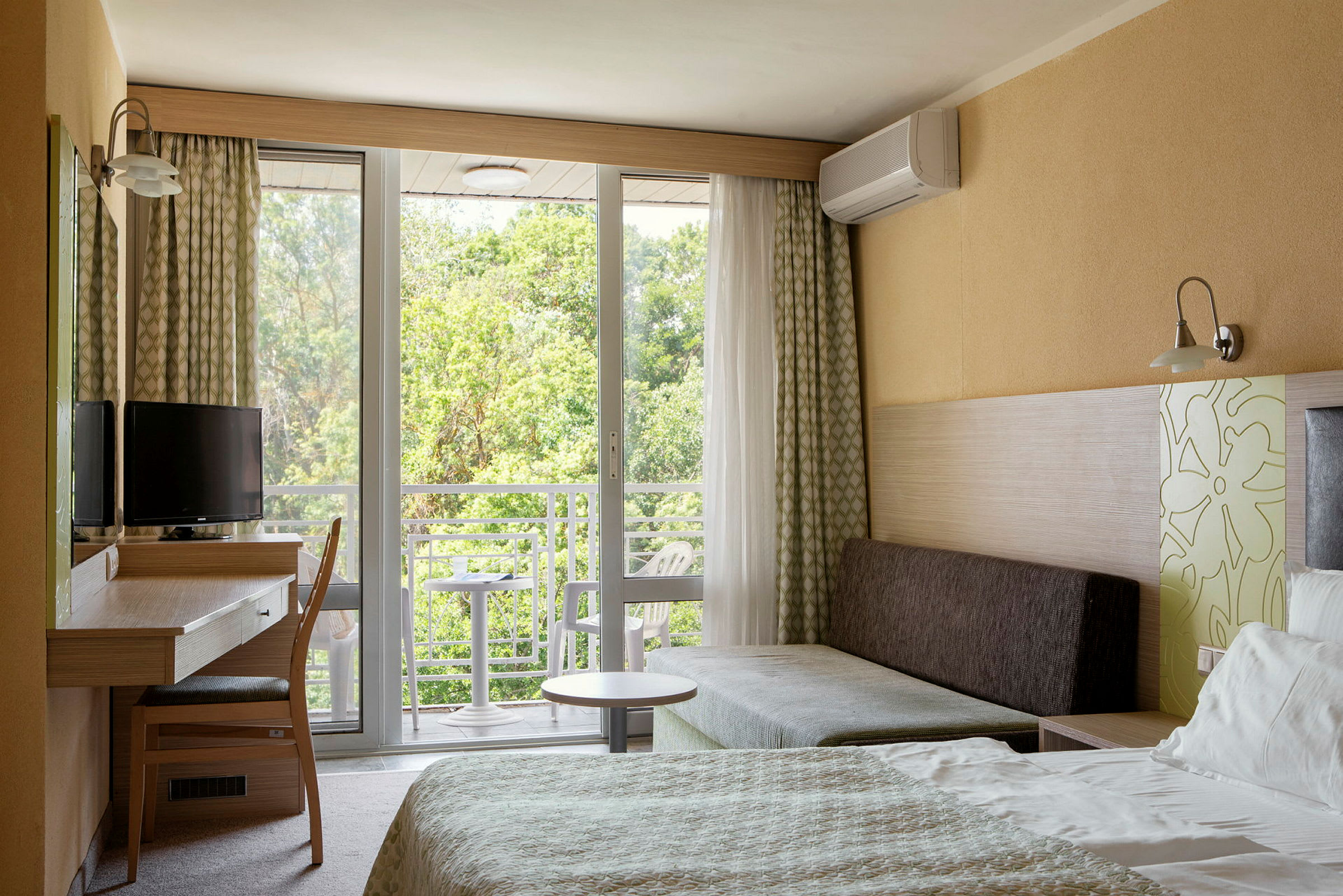 Hotellihuone, jossa on sänky, kirjoituspöytä, tuoli ja sohva. Ikkuna ja parveke, josta on näkymä vehreyteen. (Tekoälyn luoma teksti)
