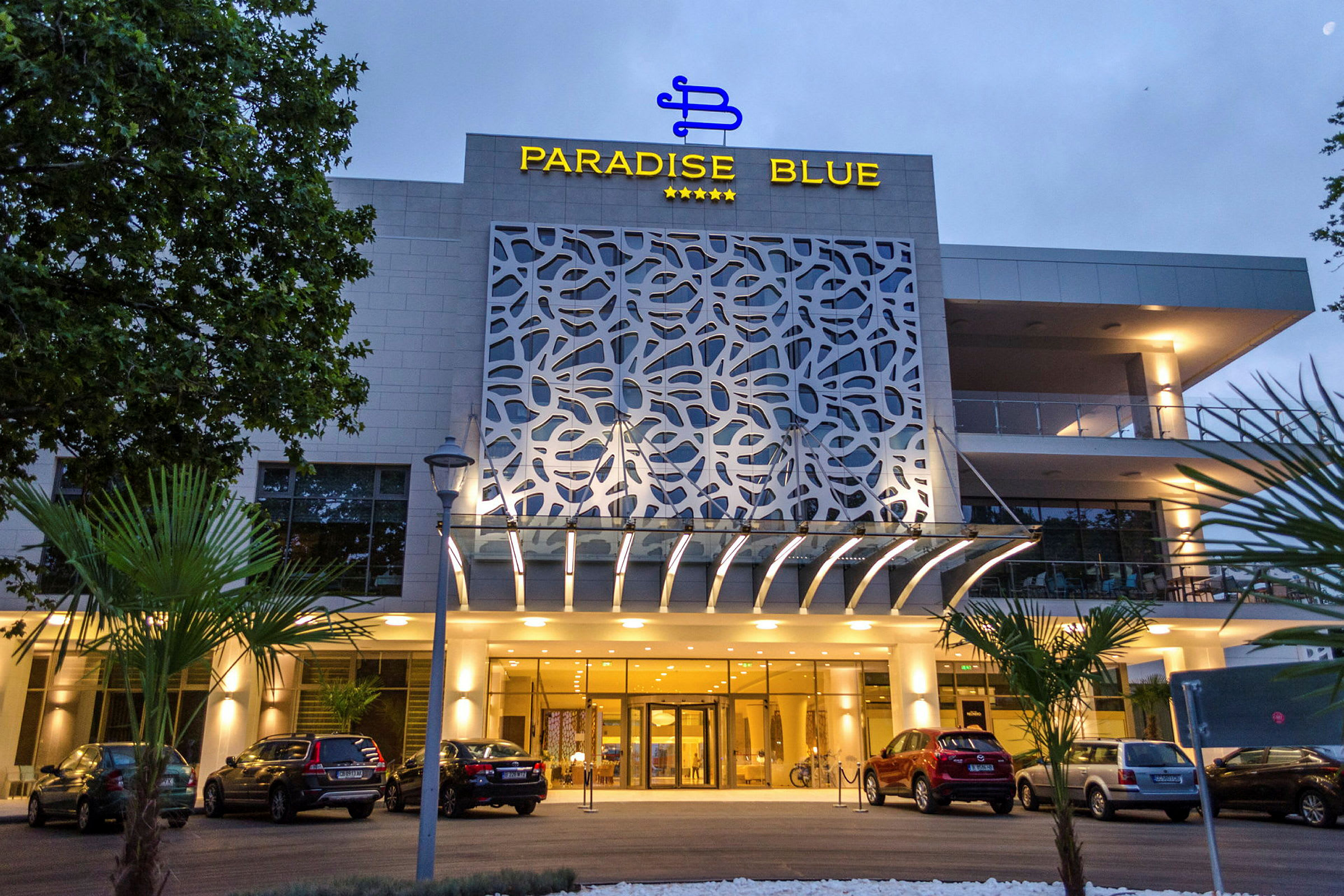 Paradise Blue -hotellin julkisivu ja sisäänkäynti autopaikoituksella. (Tekoälyn luoma teksti)