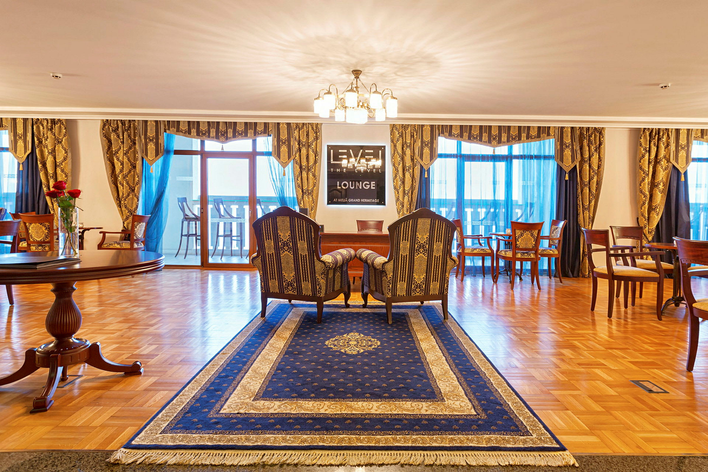 Hyvin sisustettu lounge, jossa sininen matto, pari tuolia ja pöytä, lamppuja ja koristeltuja verhoja. (Tekoälyn luoma teksti)