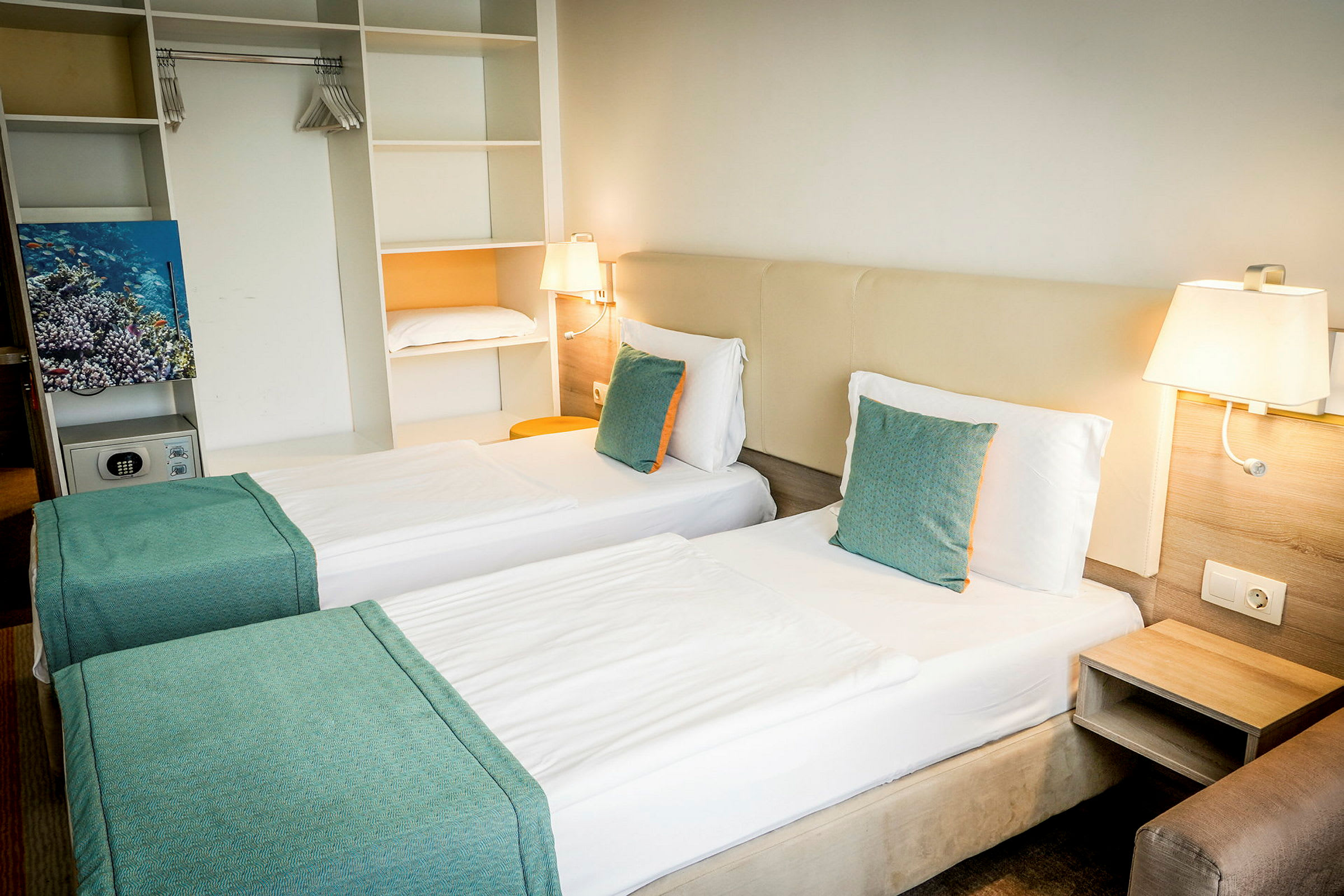 Hotellihuone, jossa kaksi yhden hengen sänkyä, valkoiset lakanat ja turkoosit tyynyt. Sisäänrakennettu säilytys ja seinävalaisimet näkyvät. (Tekoälyn luoma teksti)