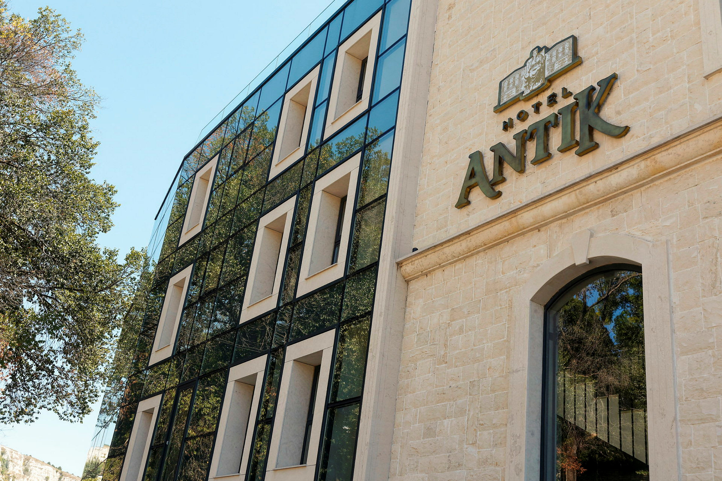 Hotellin julkisivu, jossa näkyy nimi "Hotel Antik". Modernissa rakennuksessa on suuret ikkunat ja sitä ympäröivät puut. (Tekoälyn luoma teksti)