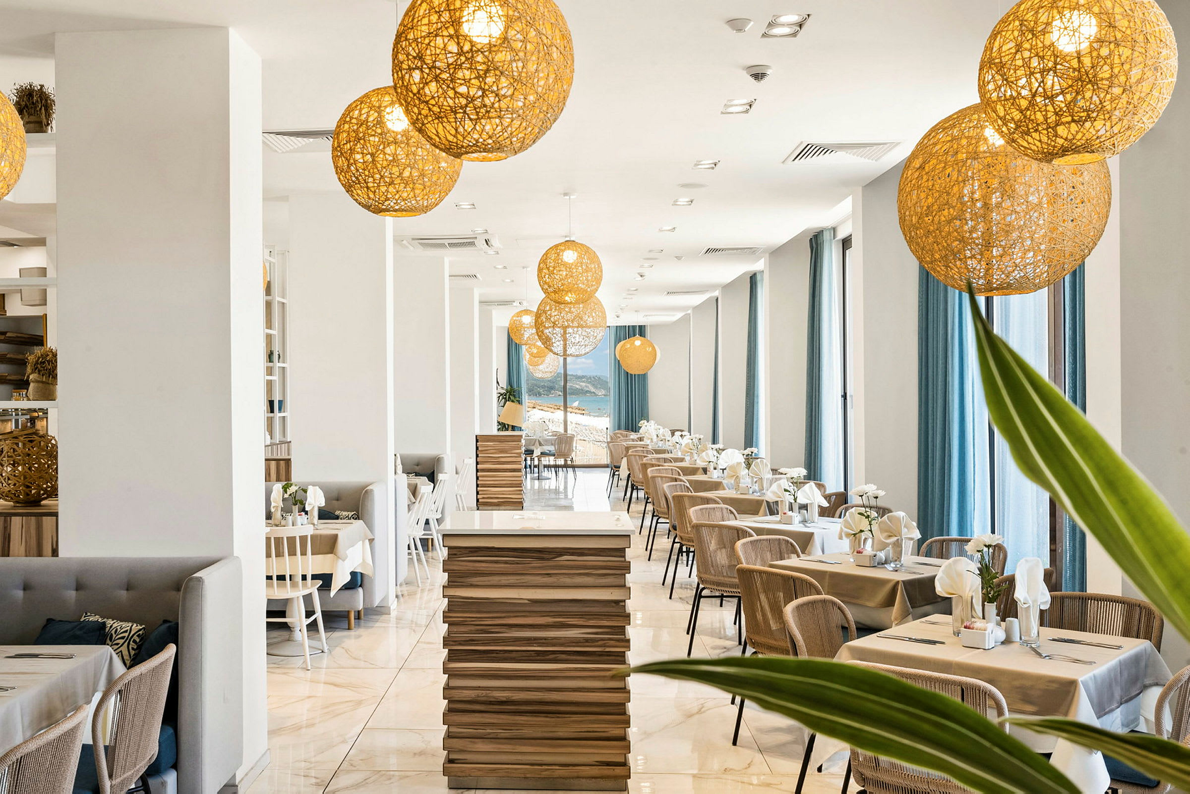 Moderni ravintola, joka on sisustettu vaaleilla puupöydillä, tuoleilla ja suurilla ikkunoilla, joissa on siniset verhot. Katosta roikkuu koristeellisia valaistusarmatuureja. (Tekoälyn luoma teksti)
