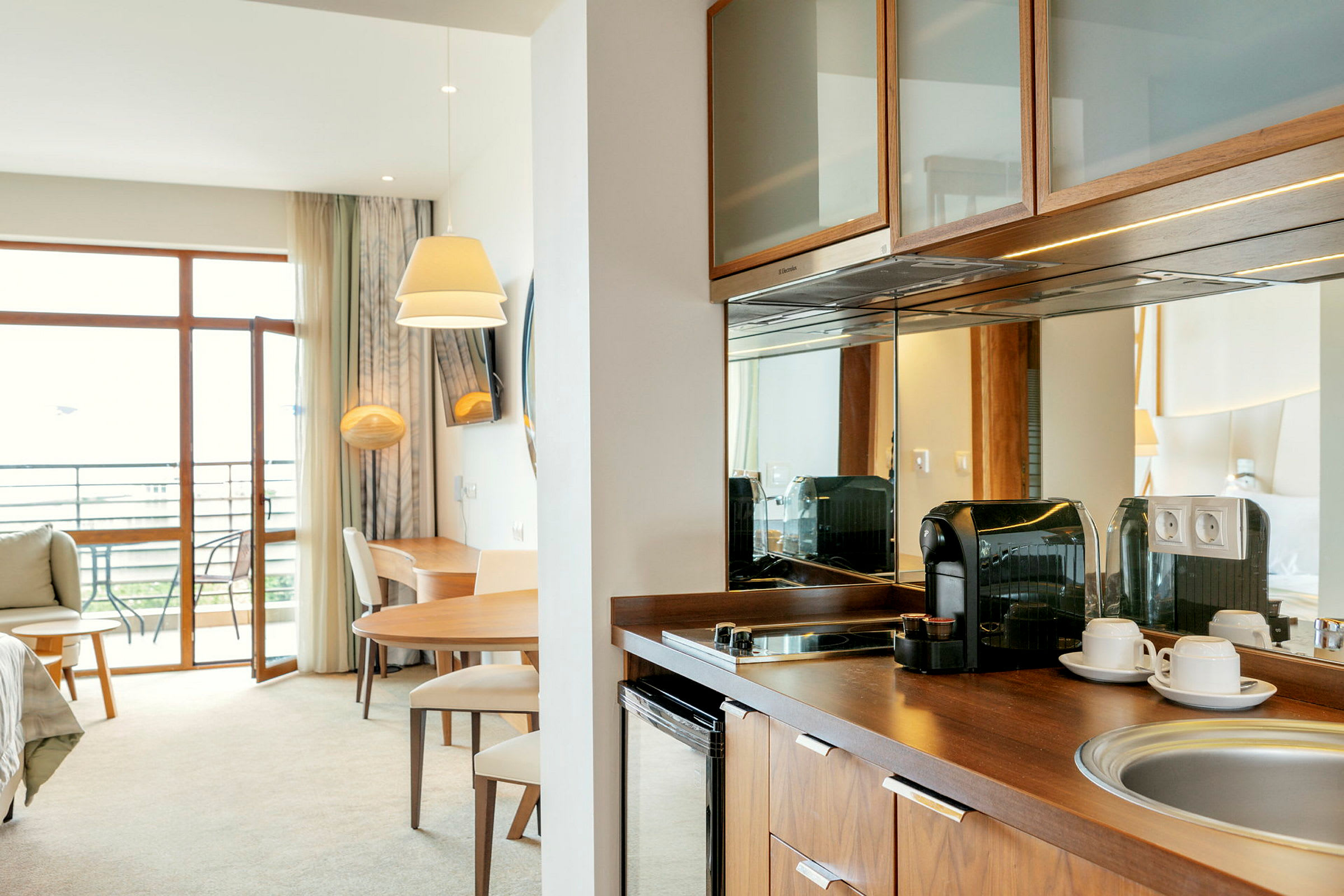 Moderni hotellihuone, jossa parveke, pöytä, tuolit, kahvinkeitin ja keittiötila. (Tekoälyn luoma teksti)