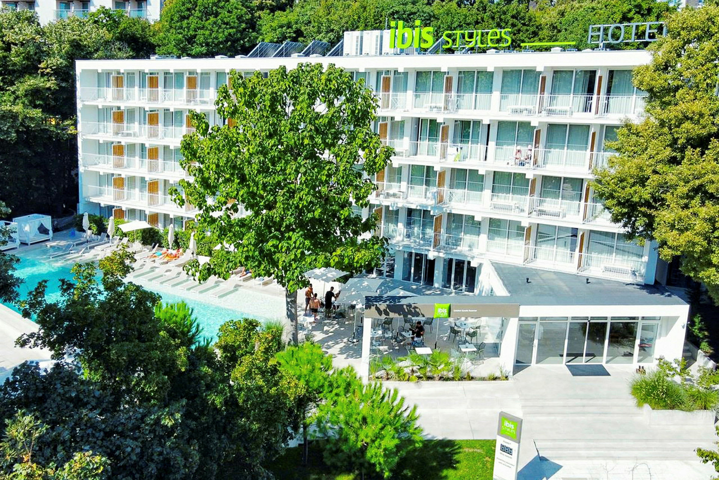 Hotellirakennus parvekkeineen uima-altaan edessä, puiden ympäröimänä. (Tekoälyn luoma teksti)