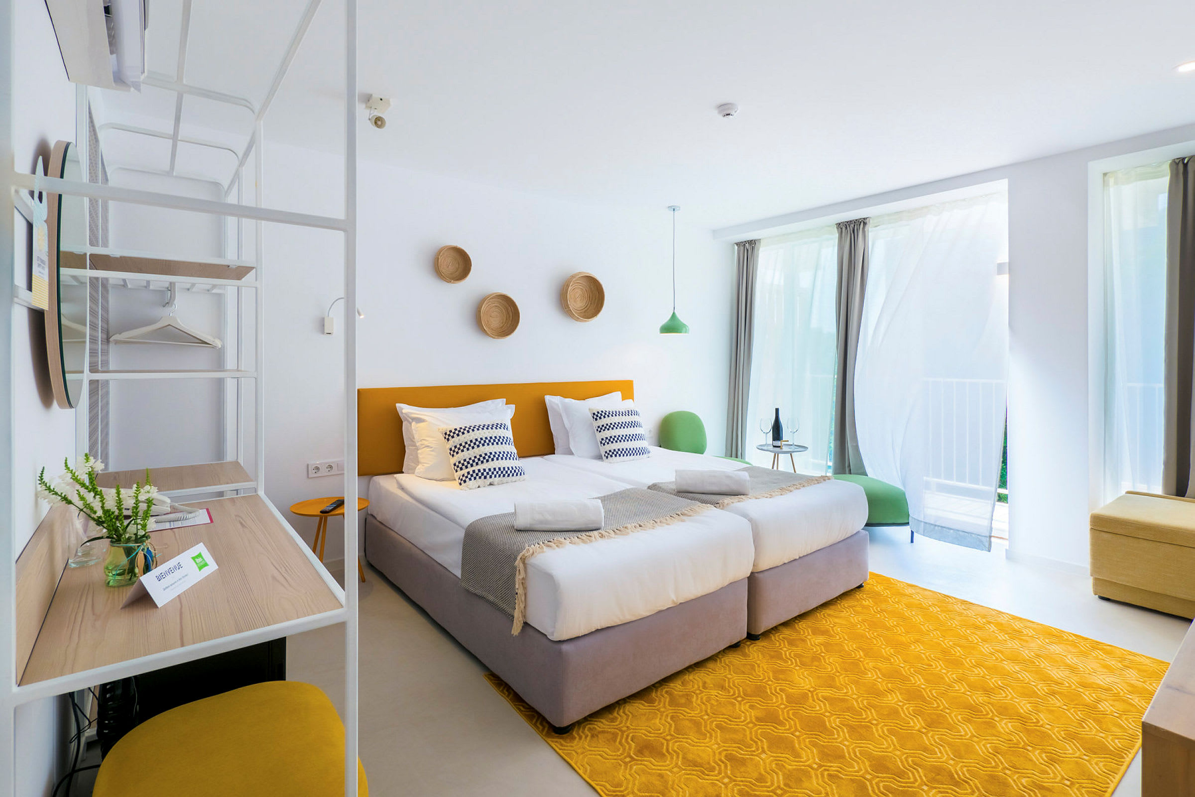 Valoisa ja moderni hotellihuone, jossa kaksi erillistä sänkyä, keltaisia yksityiskohtia ja suuret ikkunat verhoineen. (Tekoälyn luoma teksti)
