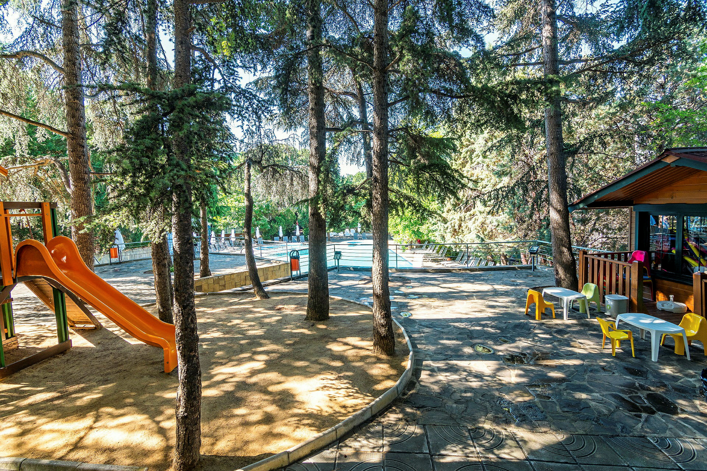 Leikkipaikka, jossa on oranssi liukumäki puiden varjossa lähellä uima-allasta. (Tekoälyn luoma teksti)