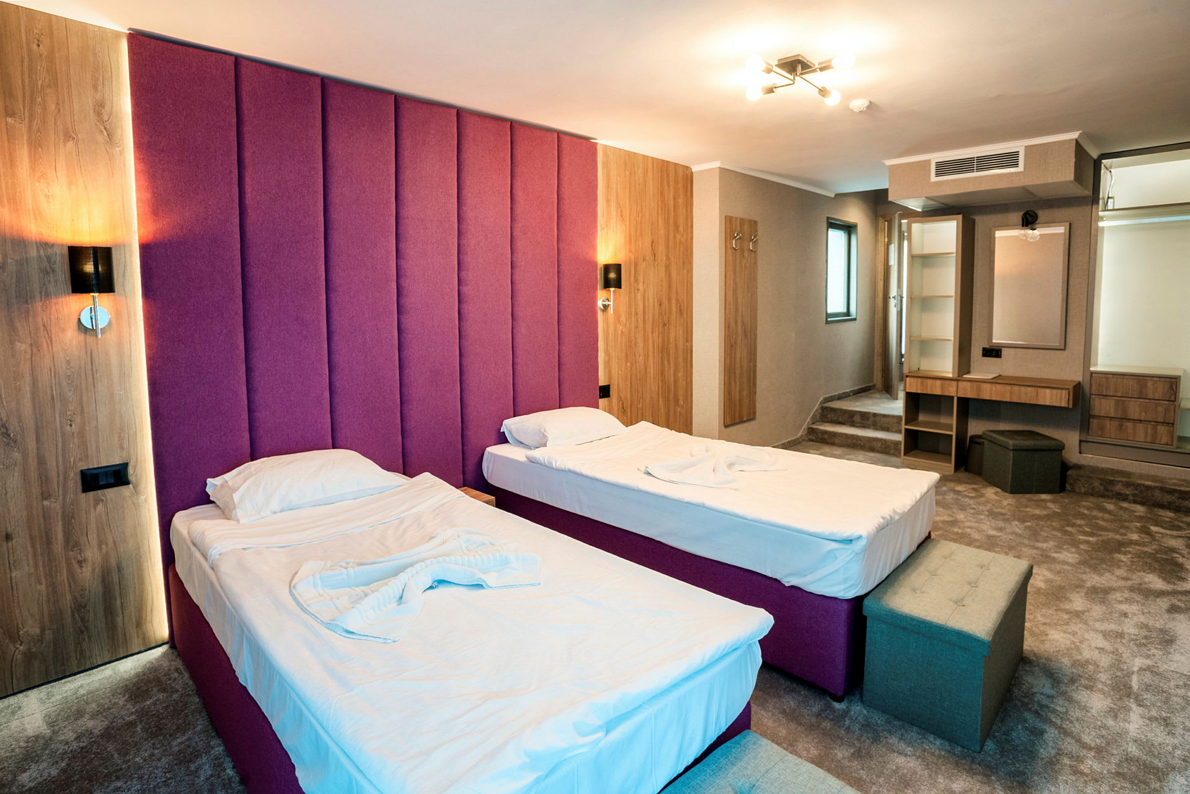 Kaksi yhden hengen sänkyä valkoisilla lakanoilla hotellihuoneessa, jossa violetti sängynpääty, puupaneelit ja työpiste peilillä. (Tekoälyn luoma teksti)