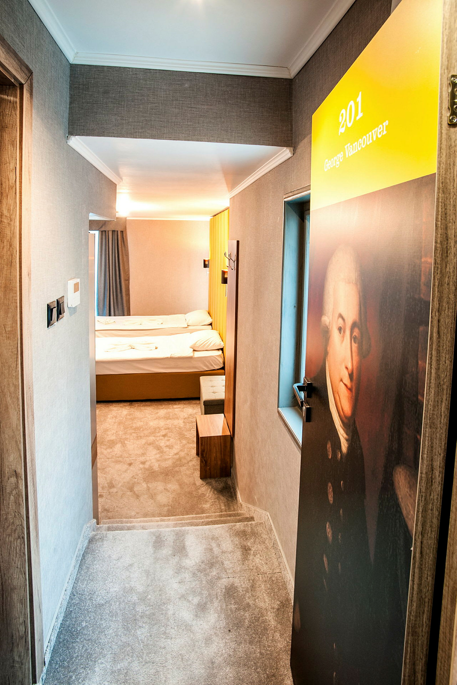 Hotellikäytävä, jossa on ovi, numeroitu 201 ja nimetty George Vancouver. Sisällä näkyy yksinkertainen hotellihuone, jossa on kaksi sänkyä. (Tekoälyn luoma teksti)