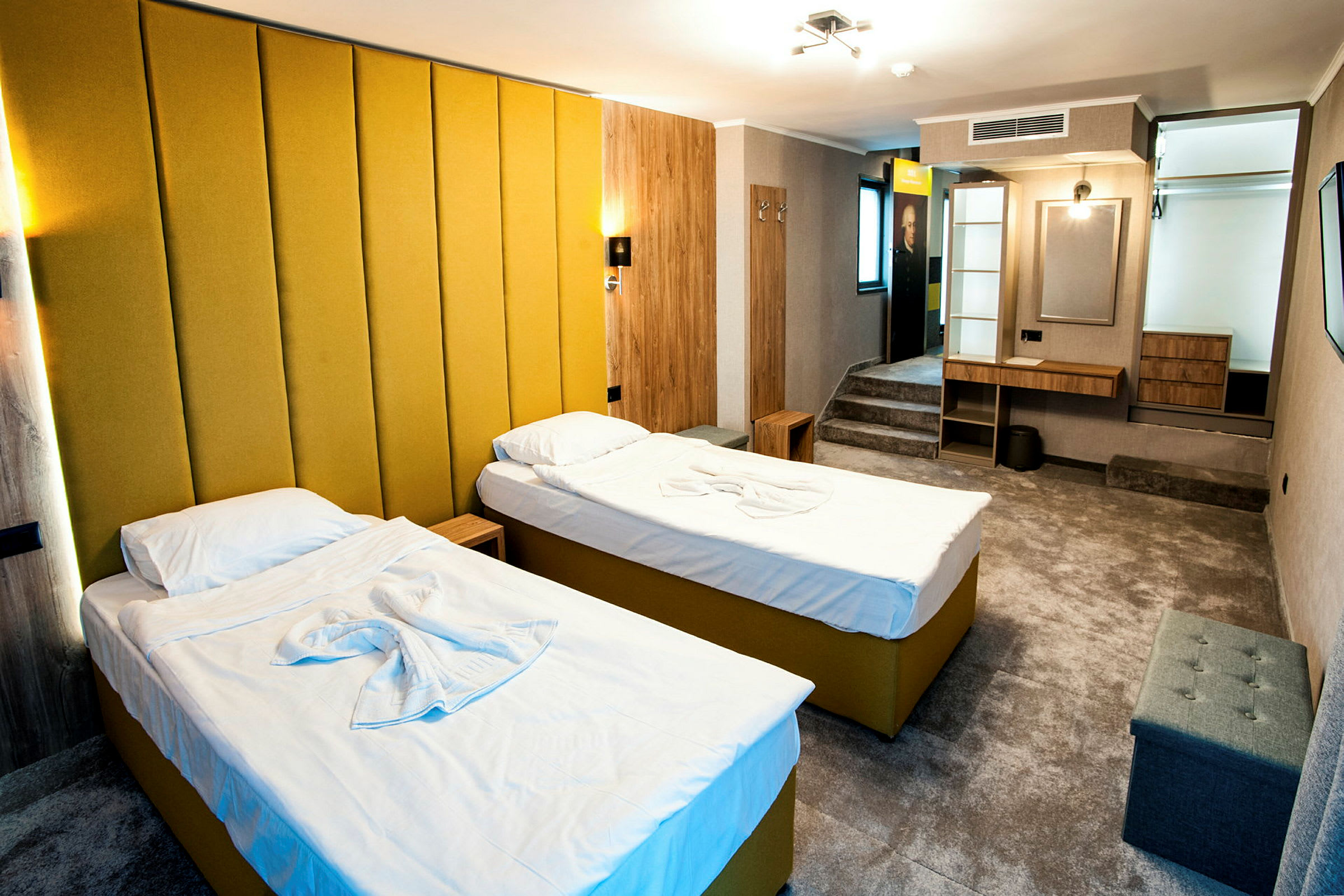 Hotellihuone, jossa on kaksi yksittäistä sänkyä keltaisilla verhoiluilla ja valkoisilla vuodevaatteilla. Huoneessa on moderni sisustus, jossa on keltainen seinäpaneeli, kirjoituspöytä, istuimet ja pieni säilytyshylly. (Tekoälyn luoma teksti)